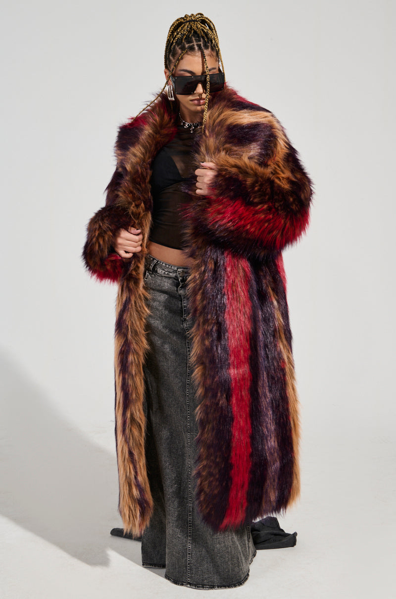 A person wearing the SUNDAY STRIPED FUR COAT, with vibrant red and purple hues, sunglasses, and a black mesh top. The oversized silhouette complements their braided hair as they stand against a plain background.