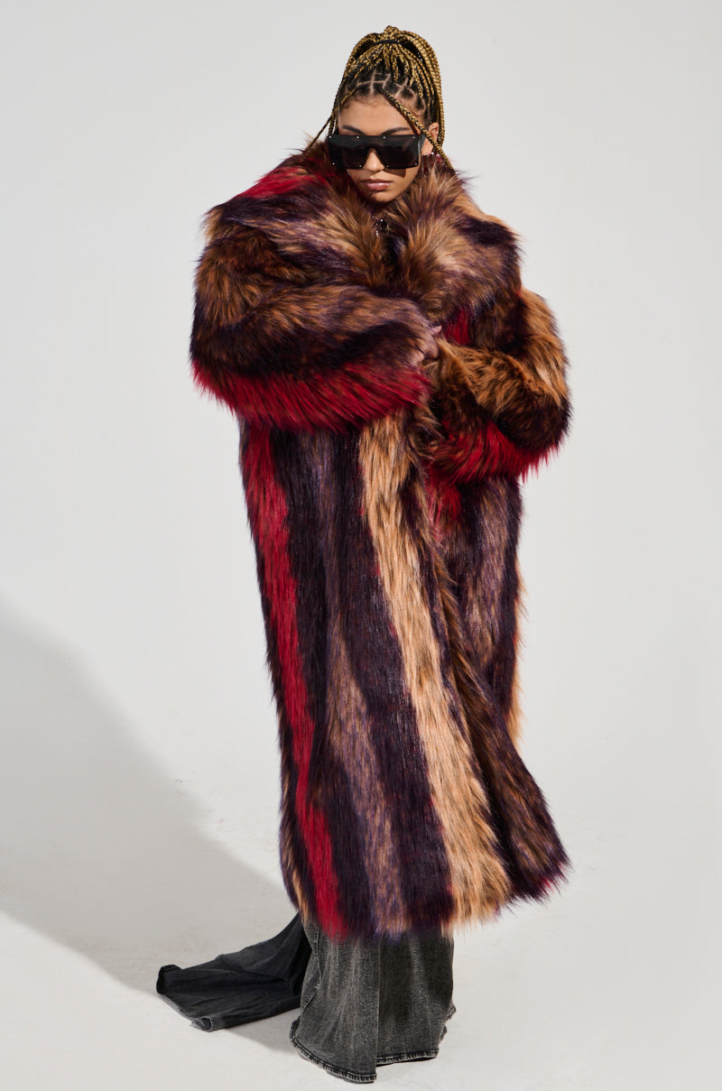 A person dressed in the SUNDAY STRIPED FUR COAT, showcasing a plush faux fur design with an oversized silhouette featuring shades of red, purple, and brown. They complement their look with dark sunglasses and have braided hair while posing against a plain white background.
