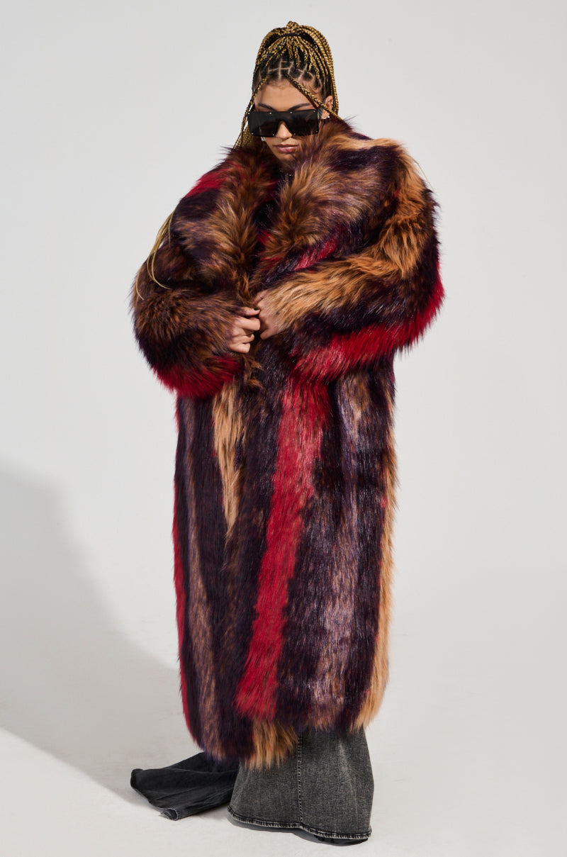 Against a simple backdrop, an individual in an oversized silhouette wears the SUNDAY STRIPED FUR COAT. The long, plush faux fur coat features red, brown, and black tones as they hold it closed. They complete the look with sunglasses and long braided hair.