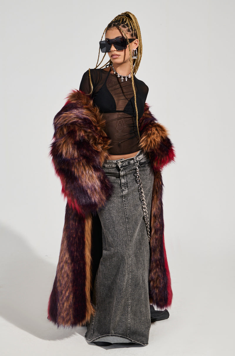 A person wearing sunglasses and the SUNDAY STRIPED FUR COAT in shades of brown and red, a sheer black top, and a long denim skirt with a chain accessory poses confidently against a plain background.