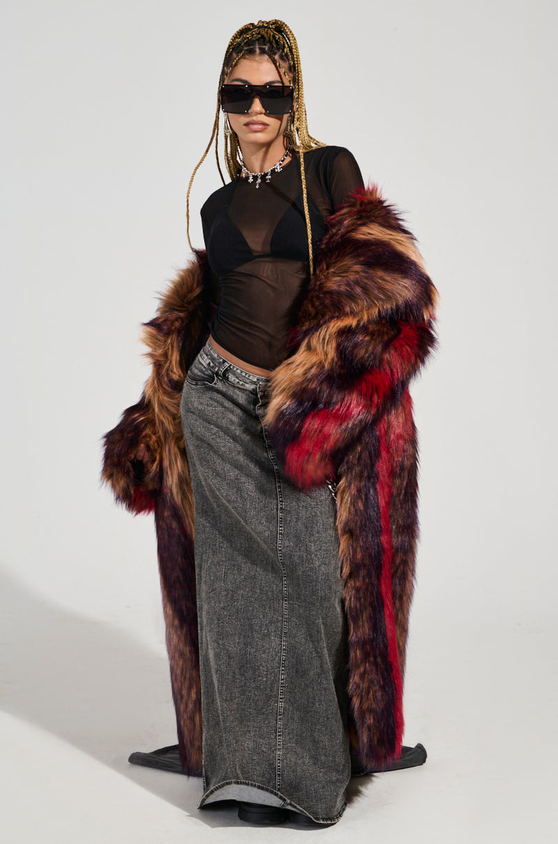 A person with elegantly styled braided hair stands confidently, showcasing a sheer black top and a long gray denim skirt. They accessorize with sunglasses, a necklace, and the oversized silhouette of the SUNDAY STRIPED FUR COAT adorned in vibrant plush faux fur featuring red and brown hues.