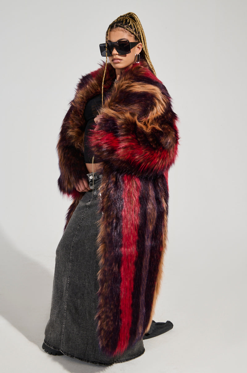 A person with braided hair confidently models the SUNDAY STRIPED FUR COAT, crafted from plush faux fur, paired with oversized sunglasses and a long dark skirt. They pose against a plain background.