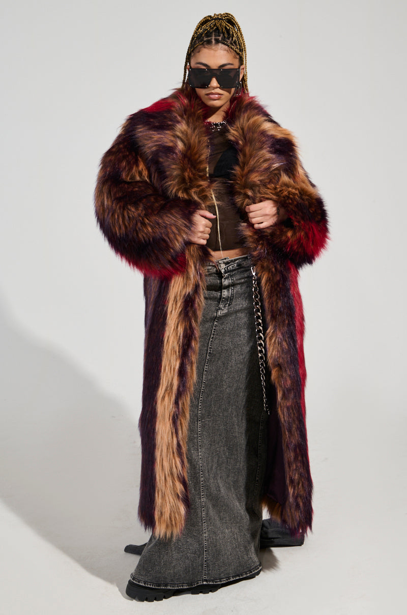 A person confidently stands wearing the SUNDAY STRIPED FUR COAT, showcasing its plush faux fur and oversized silhouette. They have braided hair, sunglasses, and a chain-linked accessory on their dark maxi skirt against a plain background.