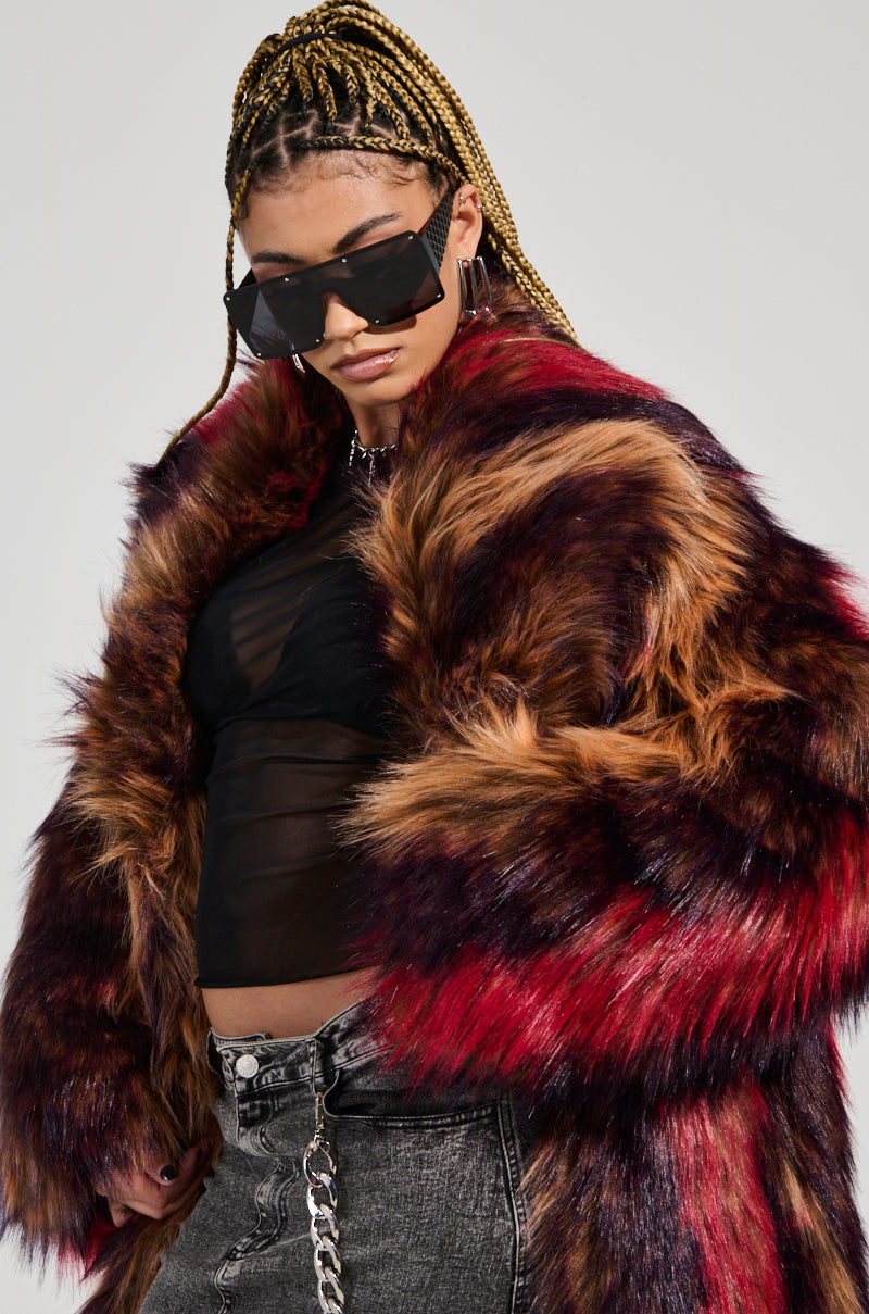 A person with braided hair wears large sunglasses and a SUNDAY STRIPED FUR COAT in shades of red and brown. They pair it with a sheer black top and gray denim jeans featuring a chain accessory, confidently embracing an oversized silhouette for a bold, fashionable look.