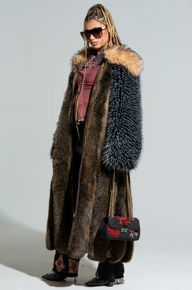 Wearing the CHIARA PANELED FUR COAT, a person shows off its various shades of brown and black faux fur. They complement the look with sunglasses, a red top, and intricately embroidered black pants. A black handbag adorned with floral designs adds a finishing touch to their chic longline silhouette. 1