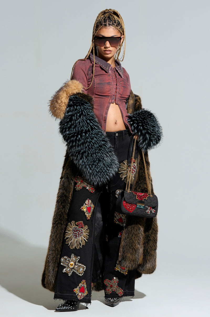 A person confidently poses wearing sunglasses, a maroon top, and the CHIARA PANELED FUR COAT. Their braided hair complements their black pants adorned with colorful embroidered designs, while a small handbag in matching design completes the ensemble. 2