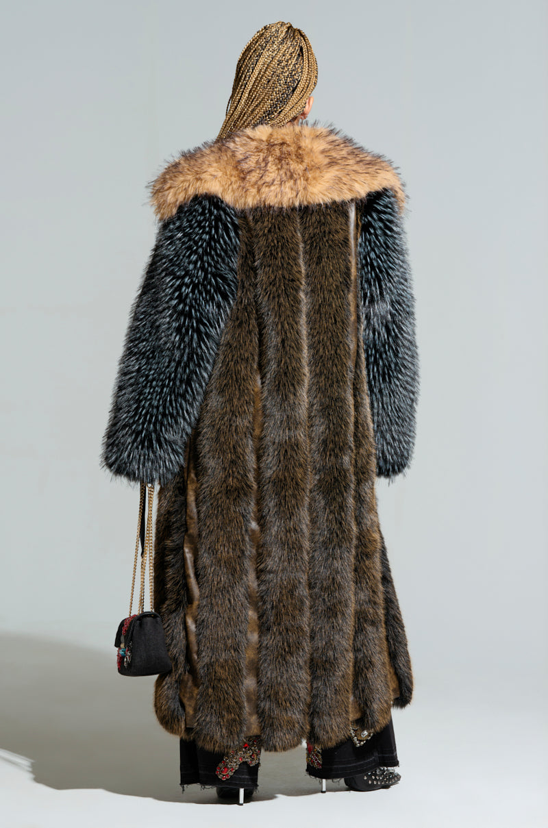 A person with braided hair is wearing the CHIARA PANELED FUR COAT, a longline silhouette made from multi-toned faux fur in a blend of brown, black, and beige. They hold a small black purse against a neutral background. 3