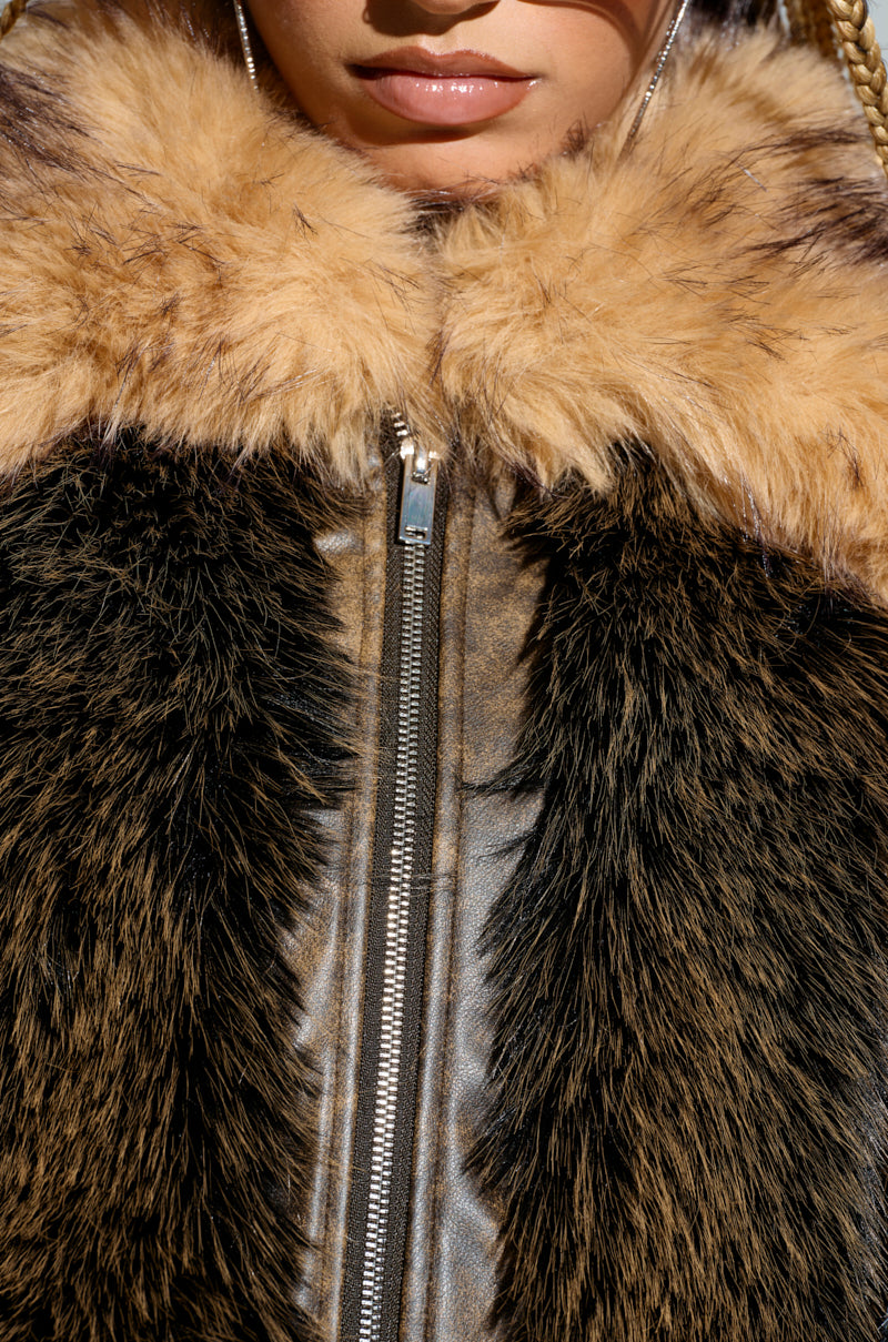 A close-up of a person wearing the CHIARA PANELED FUR COAT, featuring a longline silhouette and a shiny metallic zipper. The coat displays a combination of dark brown and light tan multi-toned faux fur panels. The person's lower face and braided hair are partially visible. 4