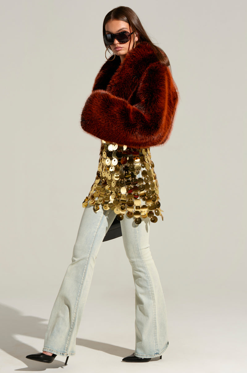 A person wearing oversized black sunglasses, the ALTER EGO FAUX FUR COAT in a cropped red style, a gold sequin mini dress, light wash flared jeans, and black pointed-toe heels strikes a pose. They stand sideways with one leg forward and arms slightly lifted against a plain, shadowed background.