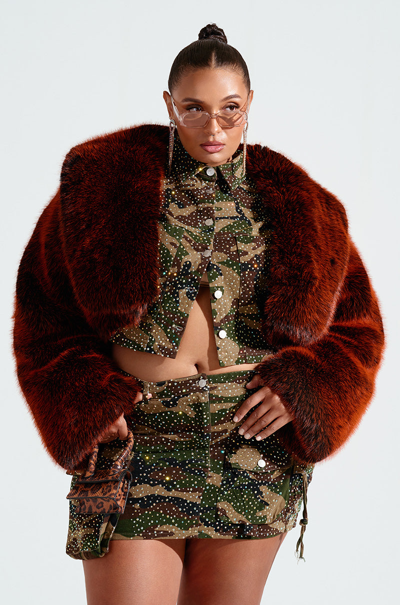 A person strikes a pose in an AKIRA Label camouflage-patterned two-piece with sparkling embellishments, complemented by the ALTER EGO FAUX FUR COAT, sunglasses, and a chic matching bag against a plain background.