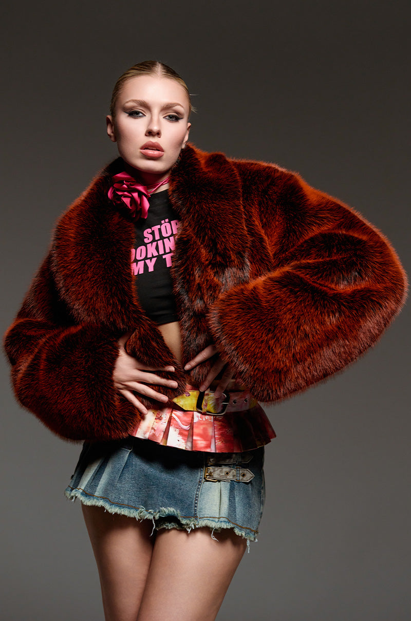 A person poses confidently against a dark background, featuring a winter ensemble highlighted by the ALTER EGO FAUX FUR COAT. They wear a black t-shirt adorned with pink lettering and pair it with a denim skirt. Their hair is neatly styled, complementing their bold, expressive look.