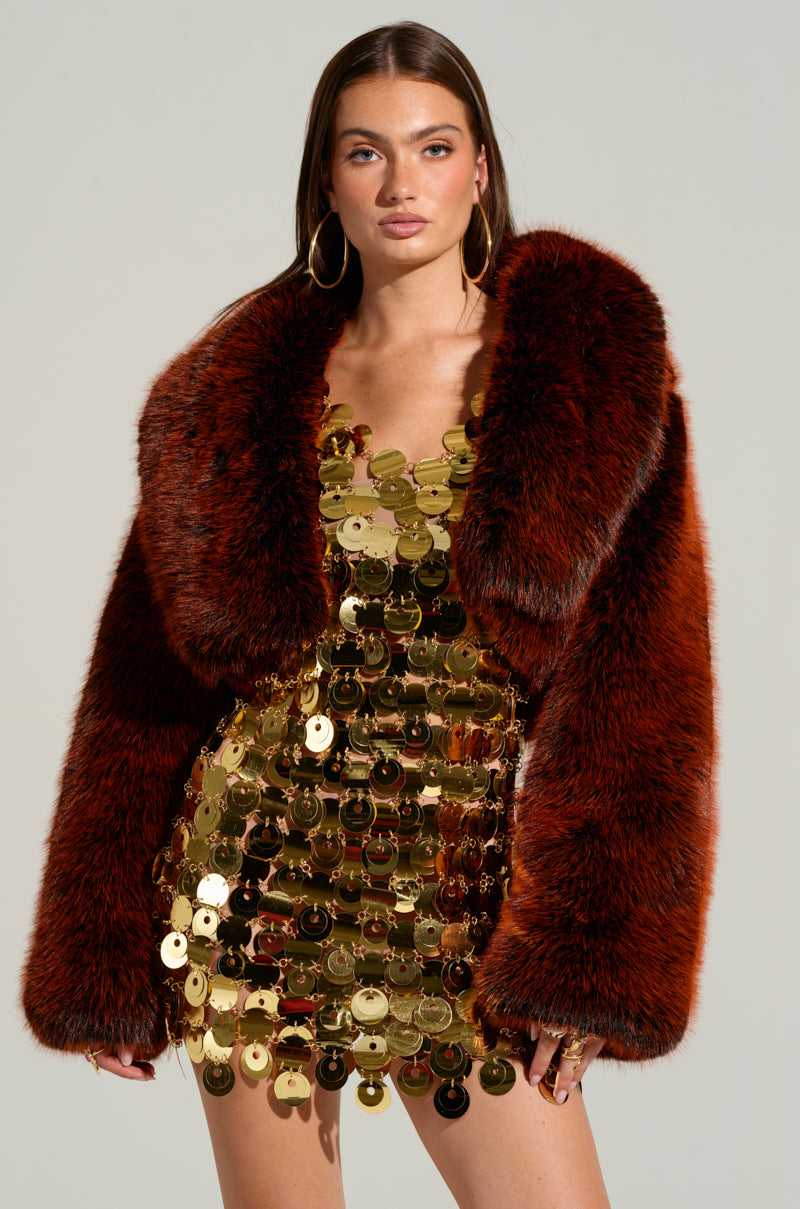 A woman poses confidently in a shiny gold sequin dress and the luxurious ALTER EGO FAUX FUR COAT. She has long brown hair, wears large hoop earrings, and stands against a plain background.