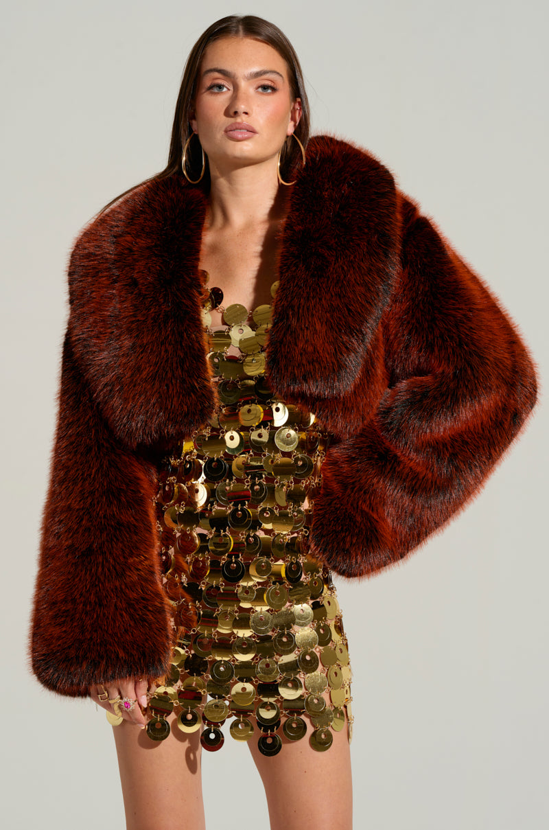 A person stands confidently in the ALTER EGO FAUX FUR COAT, an oversized dual-tone masterpiece that perfectly complements their shimmering gold sequined dress. Their long, straight hair and large hoop earrings accentuate the look against a plain background.