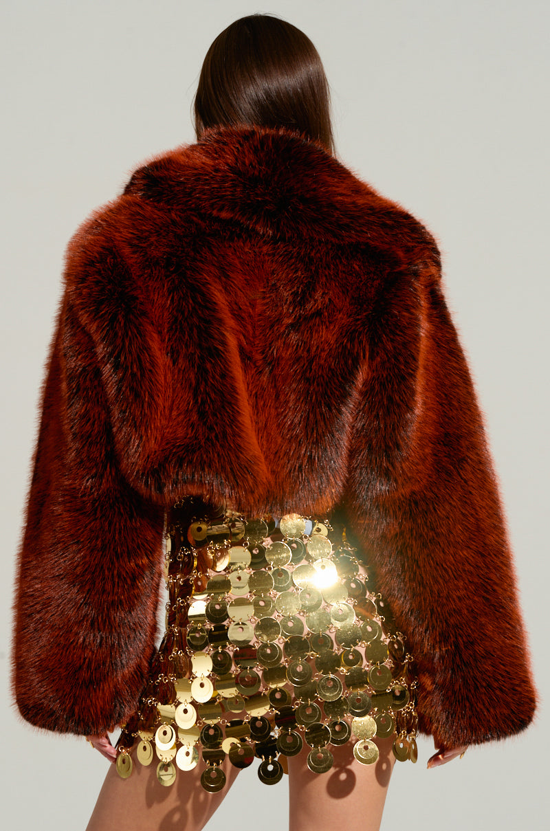 A person with long, straight hair is shown from behind wearing the ALTER EGO FAUX FUR COAT in a luxurious, deep red hue. The coat features a cropped silhouette that extends to the waist. They are also wearing a striking gold skirt adorned with large, reflective sequins. The background is a plain, light grey color.