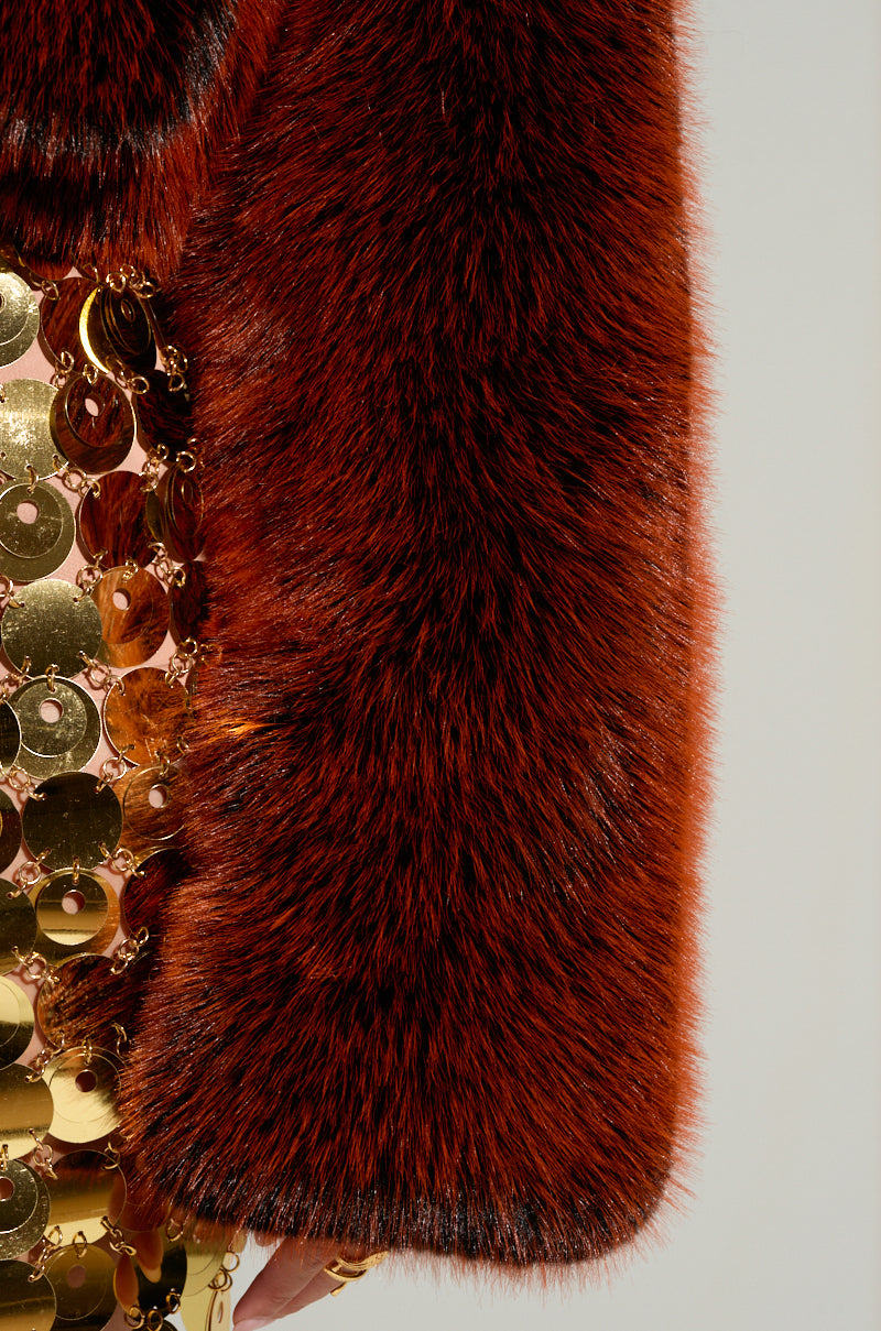 A close-up of the ALTER EGO FAUX FUR COAT showcases its luxurious design, with a reddish-brown faux fur sleeve adorned alongside a section embellished with gold, disc-shaped sequins. The contrasting textures and colors emphasize the opulence of this winter look.