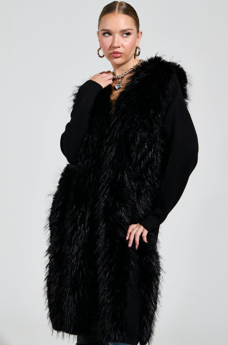 A person poses against a gray background, wearing the SASHA FIERCE LONG FUR TRIM CARDIGAN paired with hoop earrings and a chunky necklace. Their hair is slicked back, and the oversized relaxed fit of the cardigan adds an effortlessly chic vibe to the ensemble.