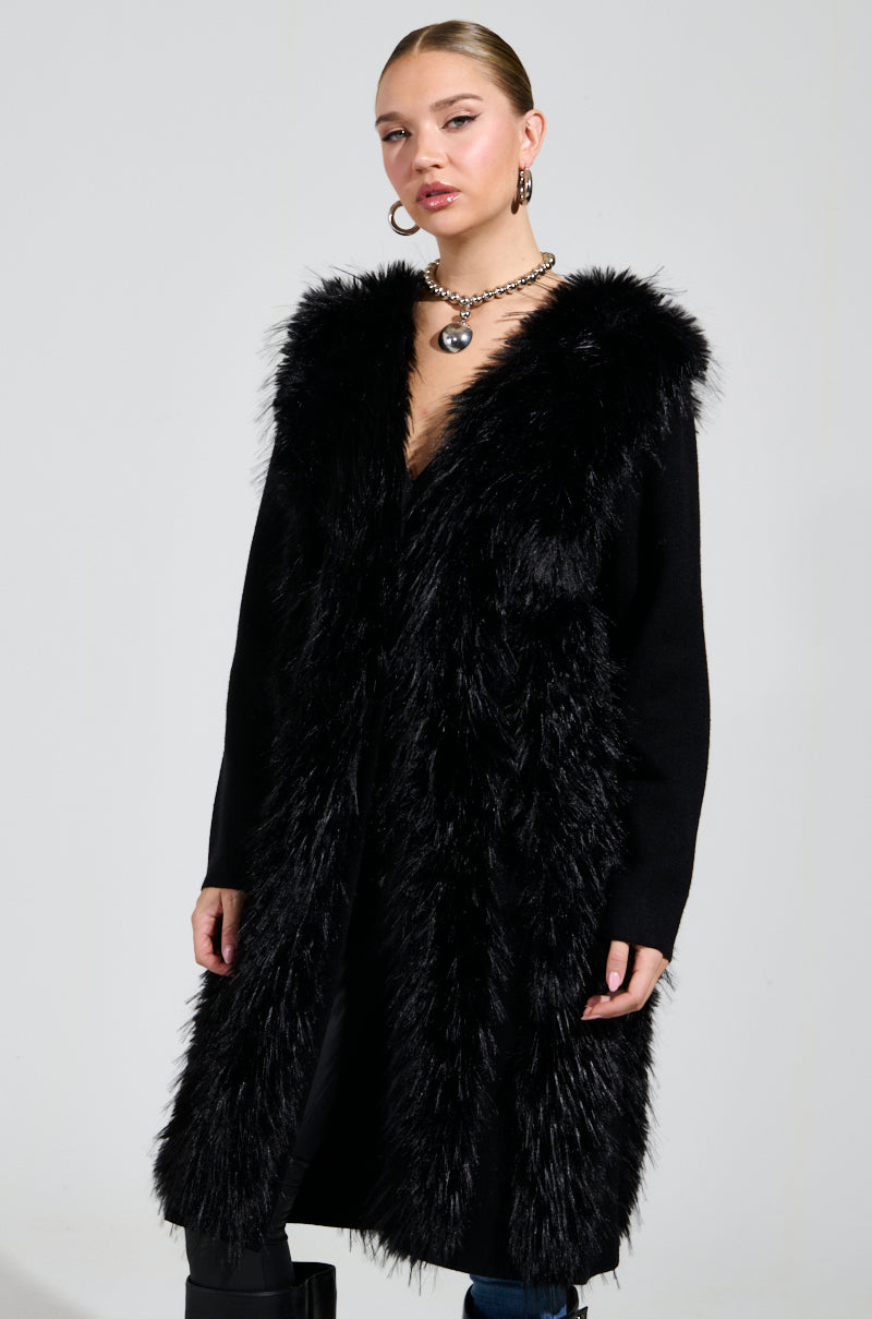 A person stands against a plain background, exuding the cozy style of an oversized relaxed fit while wearing the SASHA FIERCE LONG FUR TRIM CARDIGAN, a long black coat with a faux fur collar and deep V-neck, accessorized with a chunky necklace and hoop earrings.