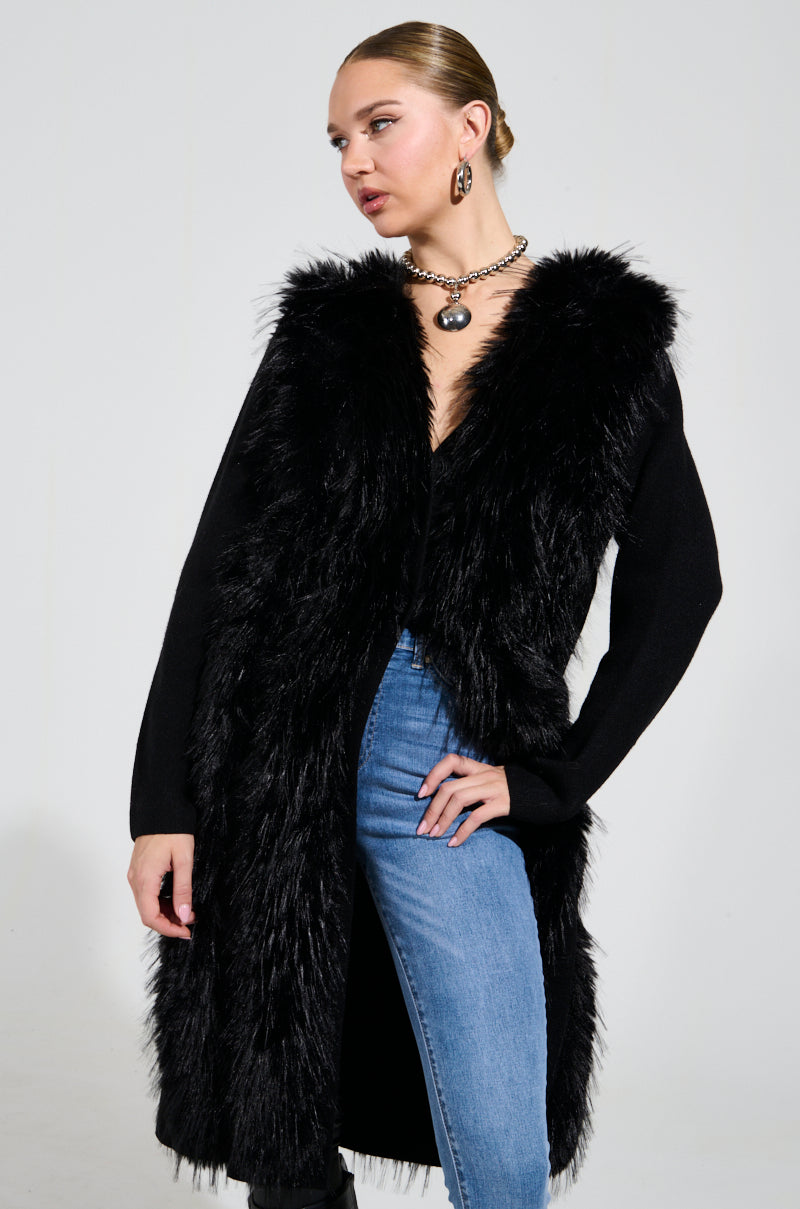 Dressed in the SASHA FIERCE LONG FUR TRIM CARDIGAN, a woman poses confidently over a black top and blue jeans. She complements her look with a chunky necklace and hoop earrings, and her hair is styled in a neat bun, exuding the elegance of an oversized relaxed-fit ensemble.