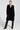 A person is standing confidently in the SASHA FIERCE LONG FUR TRIM CARDIGAN, paired with black pants and knee-high black boots. They have their hands slightly back, accessorized with hoop earrings and a necklace, against a plain white background.