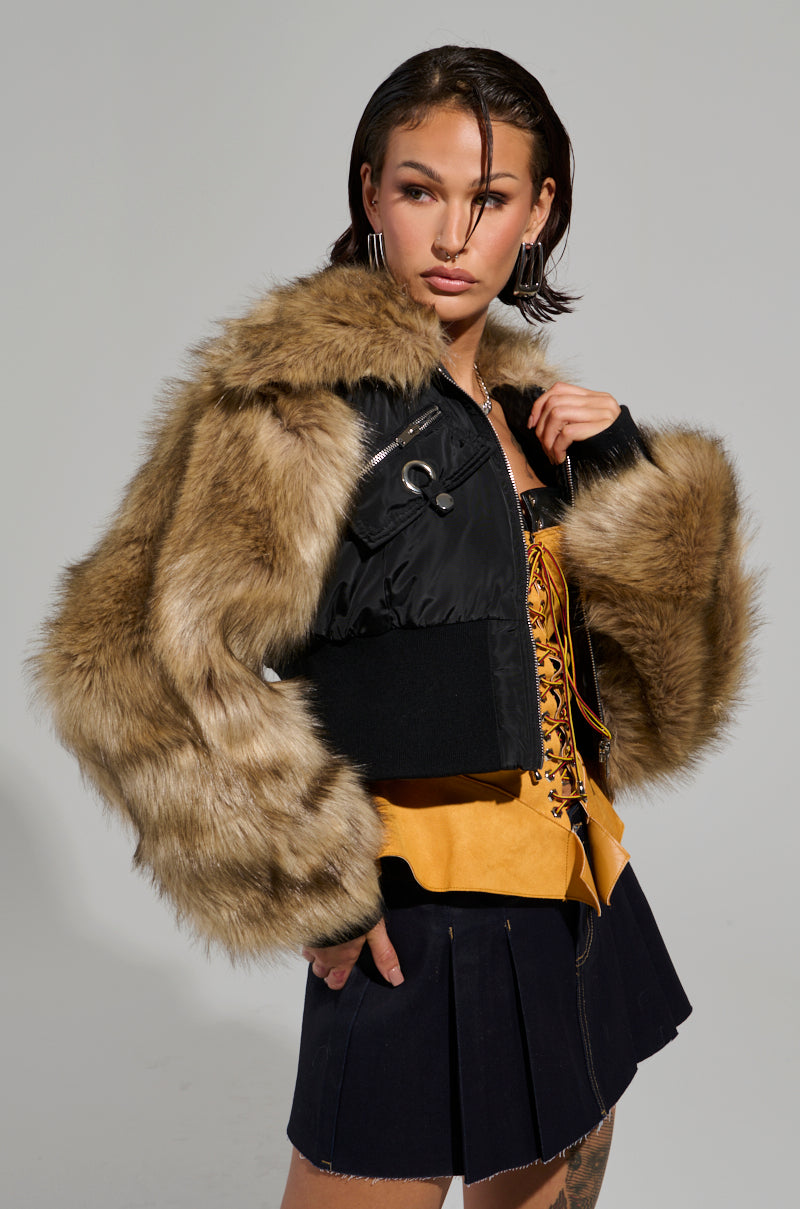 A person with short dark hair confidently poses in front of a plain background, wearing the BECKS FUR TRIM BOMBER with faux fur sleeves, a patterned corset top, and a dark pleated skirt.
