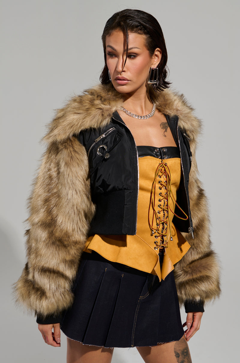 A person wearing the BECKS FUR TRIM BOMBER with quilted inner lining and a yellow lace-up top stands against a plain background. The outfit includes a pleated skirt and a chain necklace. The individual has short, styled hair and exudes confidence.