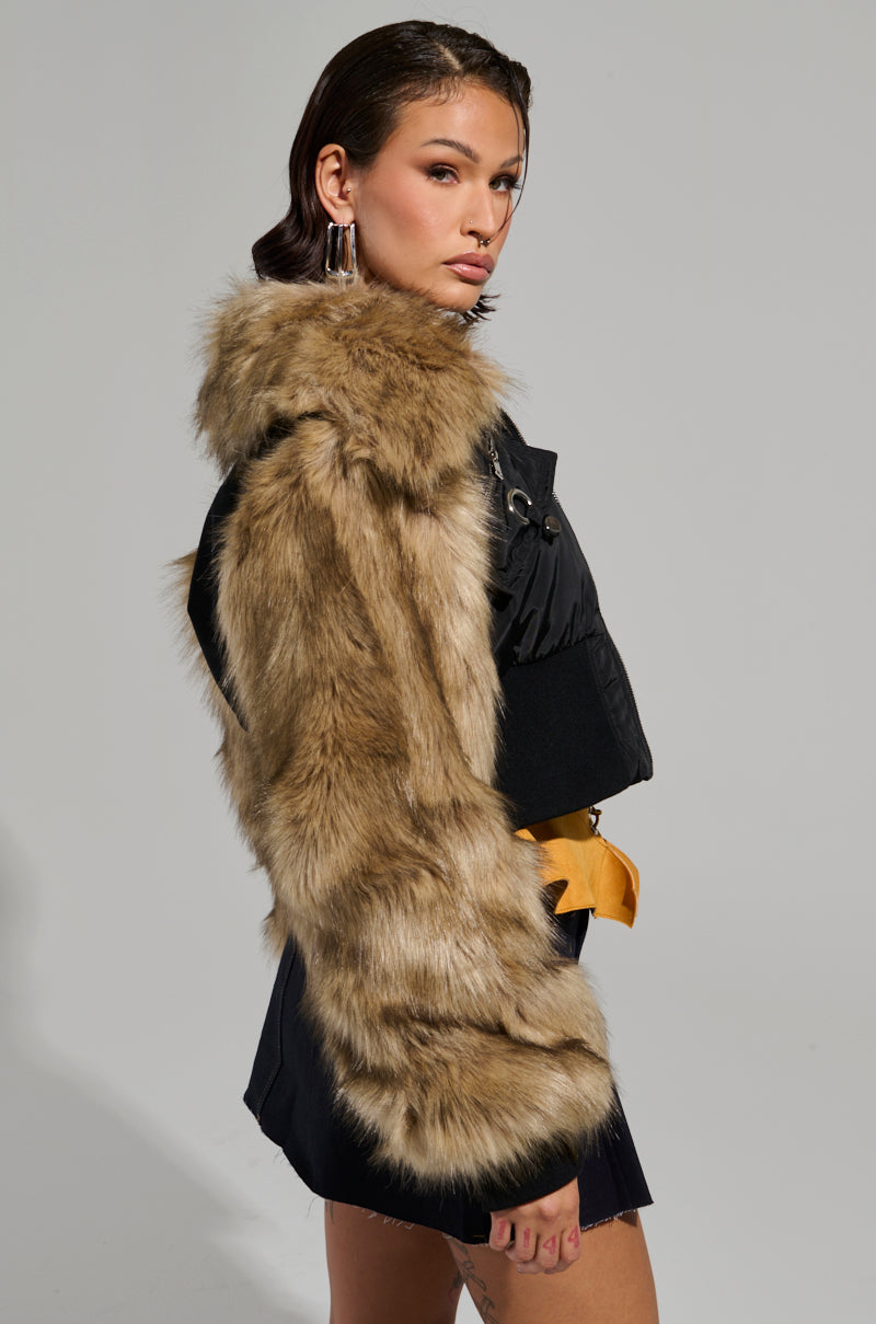 A woman poses confidently in the BECKS FUR TRIM BOMBER, a sleek black outfit with faux fur sleeves and a quilted inner lining. Her shoulder-length hair and large hoop earrings add flair as she stands against a plain backdrop.