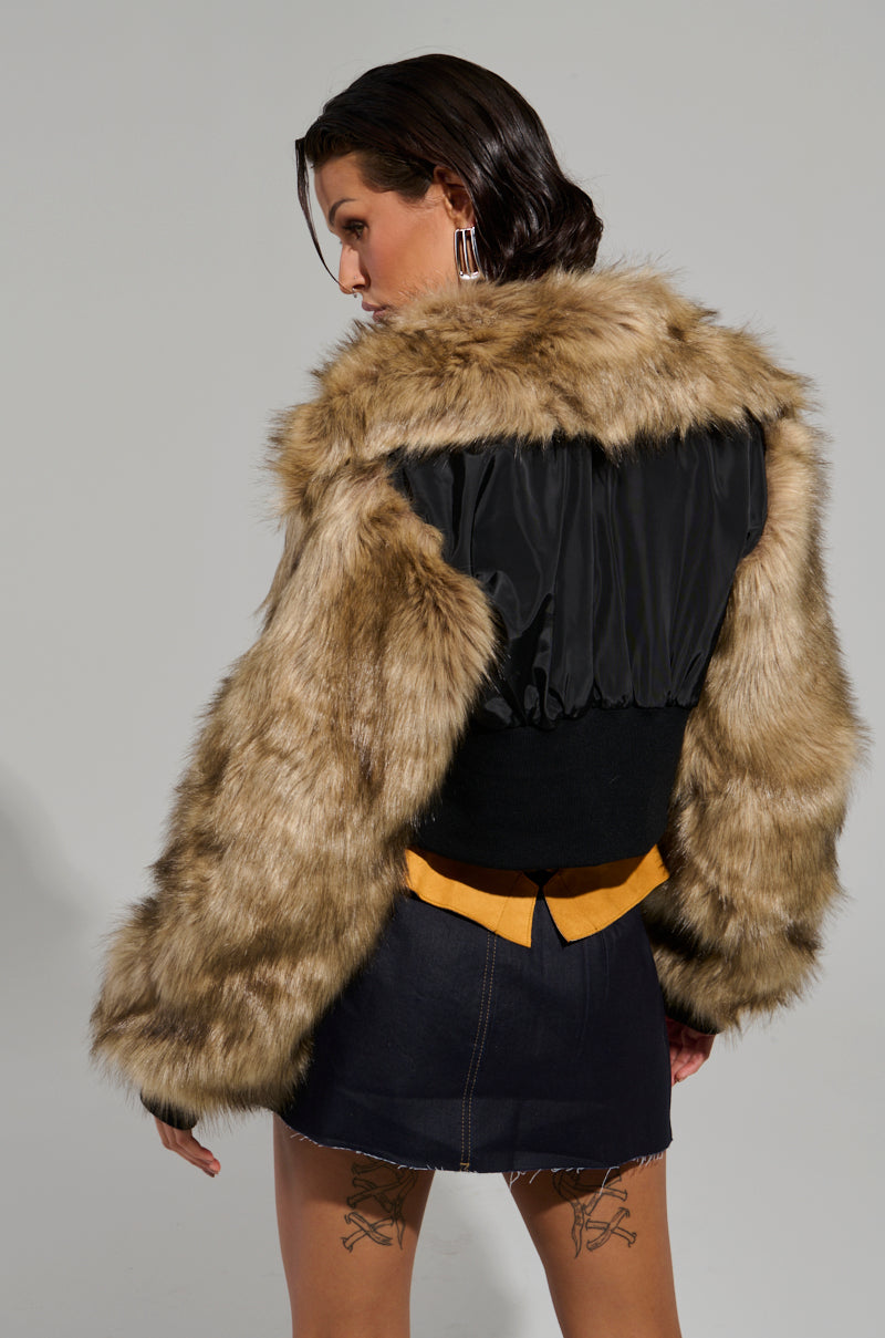 A person with dark wet hair is slightly turned, wearing the BECKS FUR TRIM BOMBER featuring a faux fur jacket with quilted inner lining. They pair it with a black top and a dark denim skirt that reveals visible tattoos on their thighs, standing against a simple backdrop.