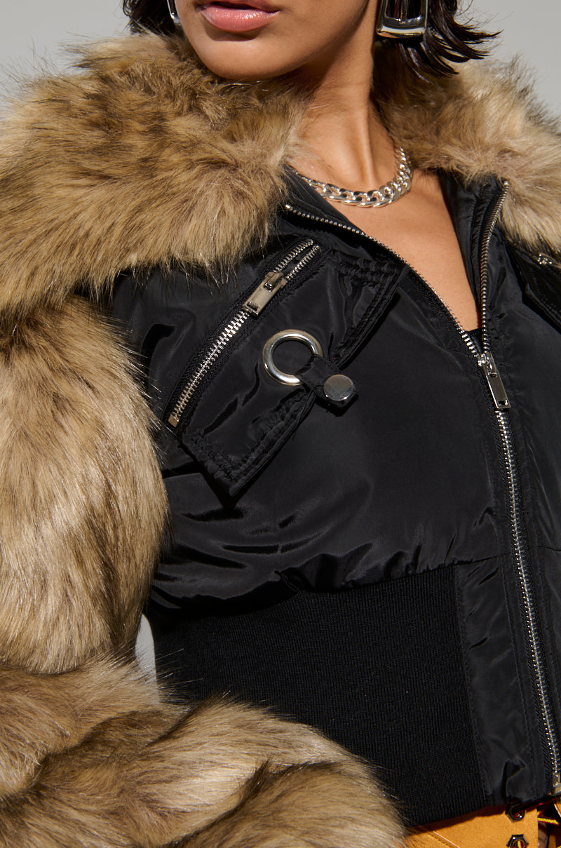 The person is wearing the BECKS FUR TRIM BOMBER, a stylish black bomber jacket with quilted inner lining, featuring multiple zippers and a ring detail. A thick chain necklace and hoop earrings complement the chic ensemble, adding flair to the outfit.