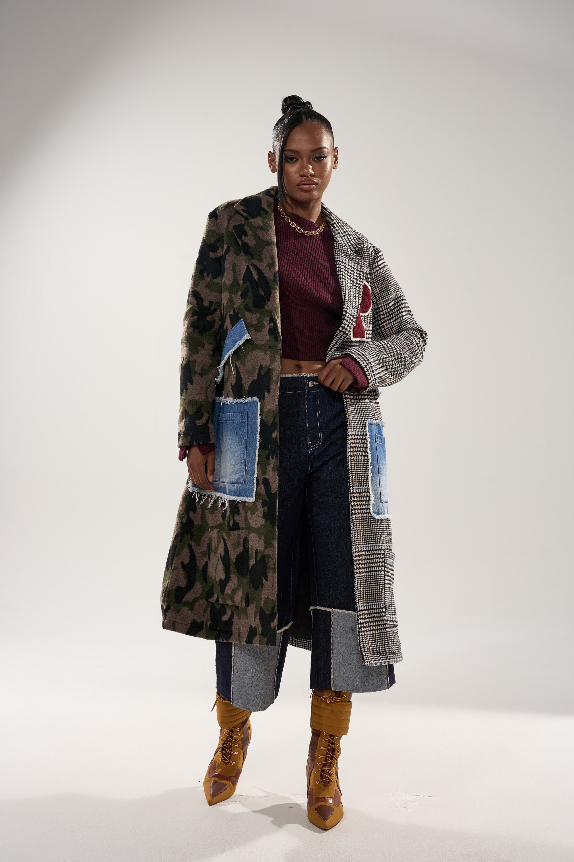 A woman stands confidently in the MADDIX HALF HALF TRENCH, featuring camo and plaid accents, paired with a maroon crop top, wide-leg cuffed jeans with blue patches, mustard lace-up boots, and gold jewelry.