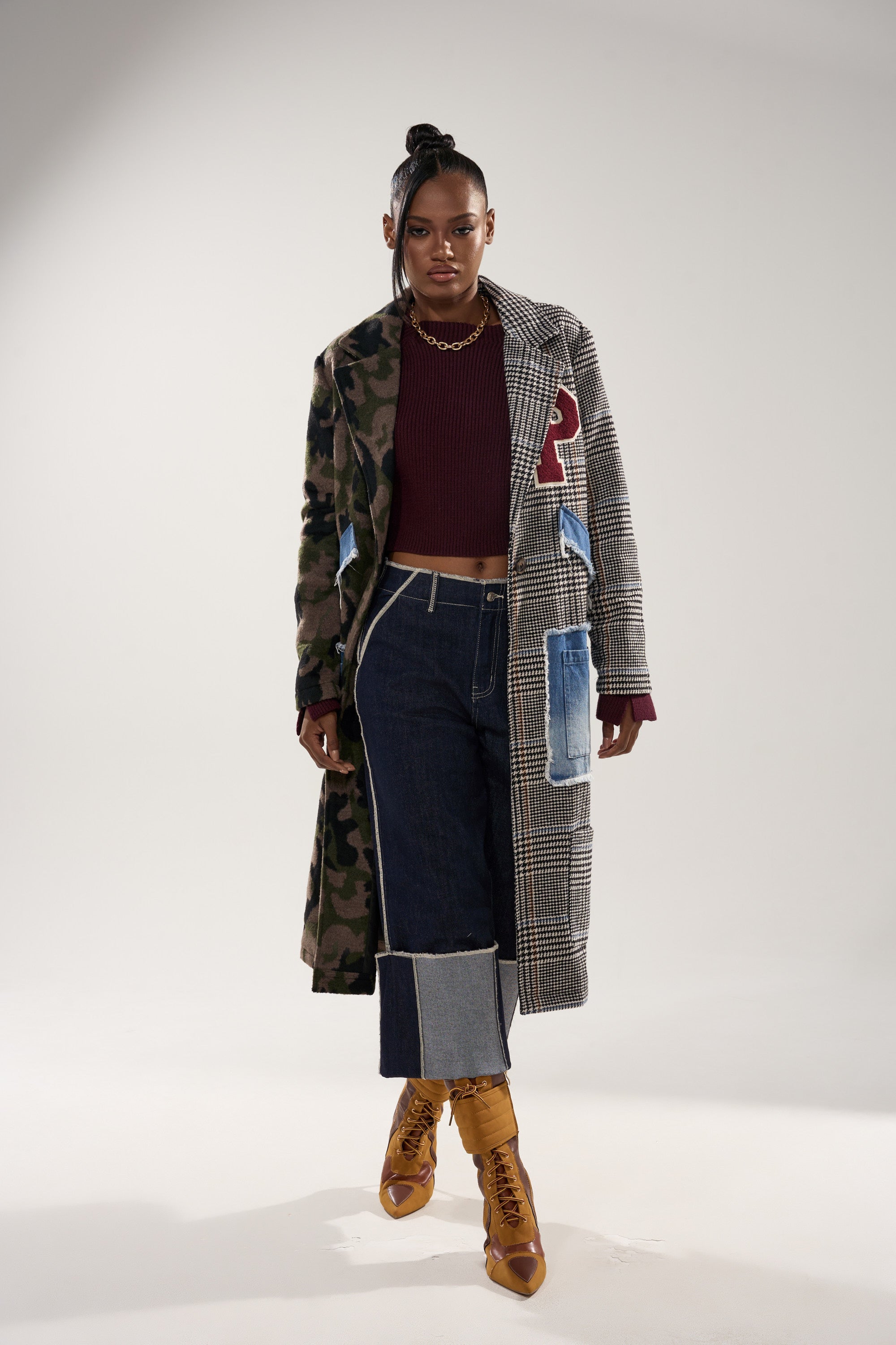 A woman stands confidently in the MADDIX HALF HALF TRENCH with patchwork patterns, a cropped maroon sweater, wide-leg dark jeans with light blue cuffs, tan lace-up boots, and a gold chain necklace against a plain background.