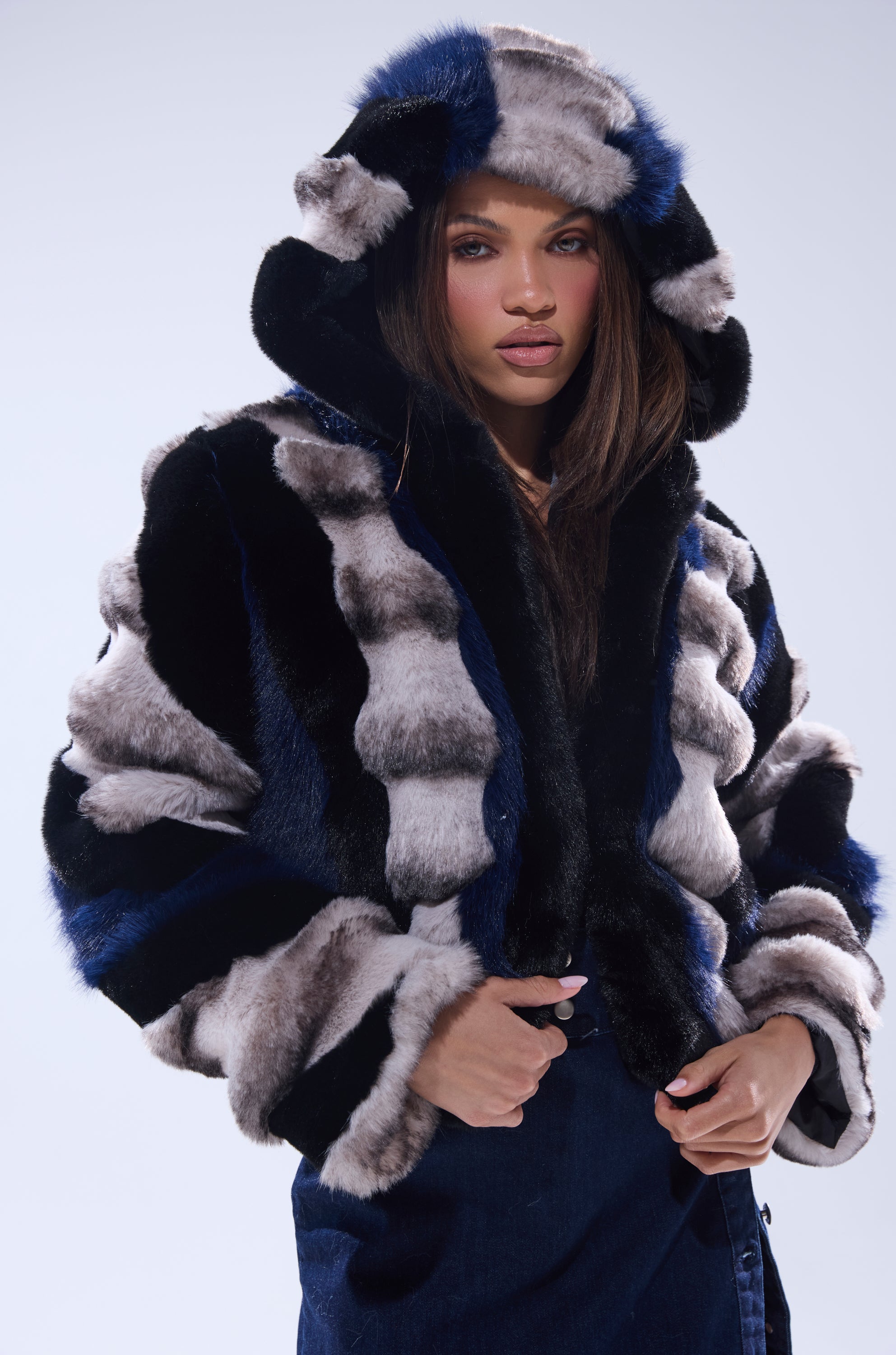 A woman models the SUPERNOVA PANELED FAUX FUR IN BLUE MULTI, a hooded coat with bold black, gray, and blue stripes, standing against a light background and looking directly at the camera.