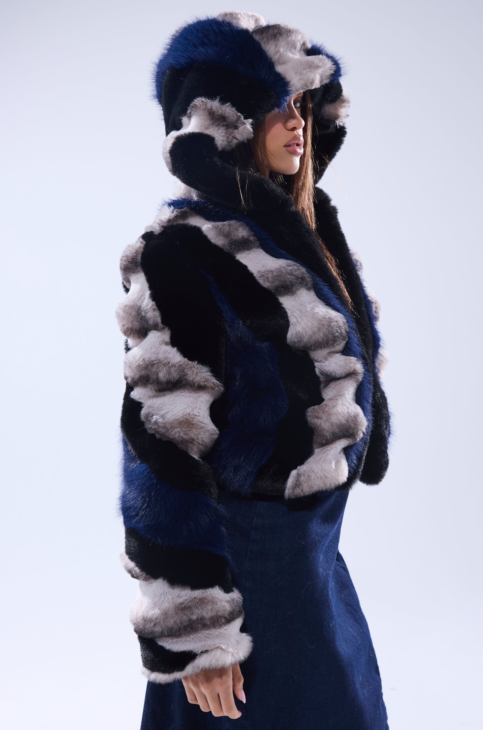 A person wearing the SUPERNOVA PANELED FAUX FUR IN BLUE MULTI, featuring bold blue, black, and white stripes and a hood, stands in profile against a plain light background.