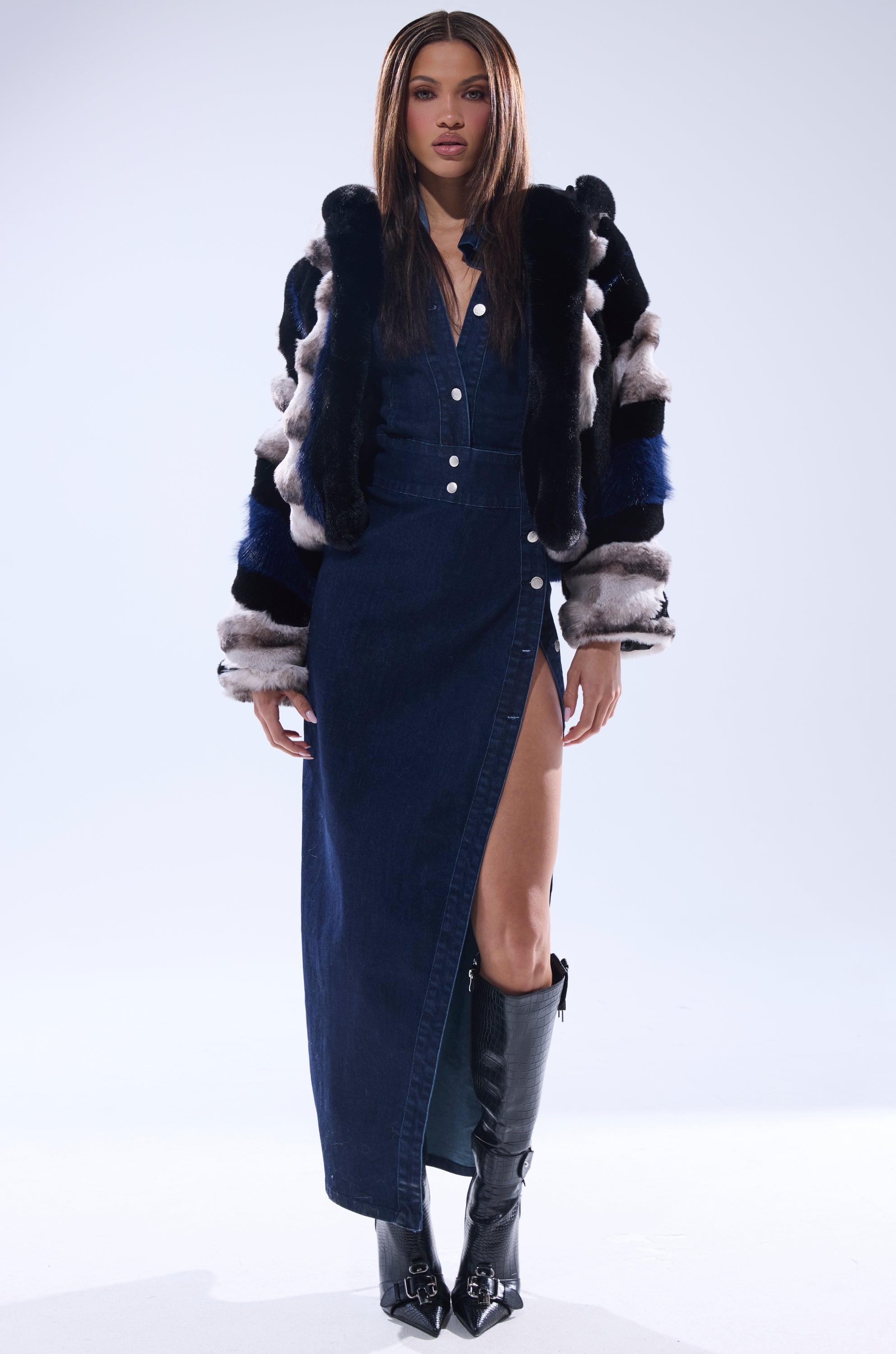 A woman stands confidently in a dark denim button-up dress with a high slit, styling it with the SUPERNOVA PANELED FAUX FUR IN BLUE MULTI jacket and knee-high black boots against a plain light background.