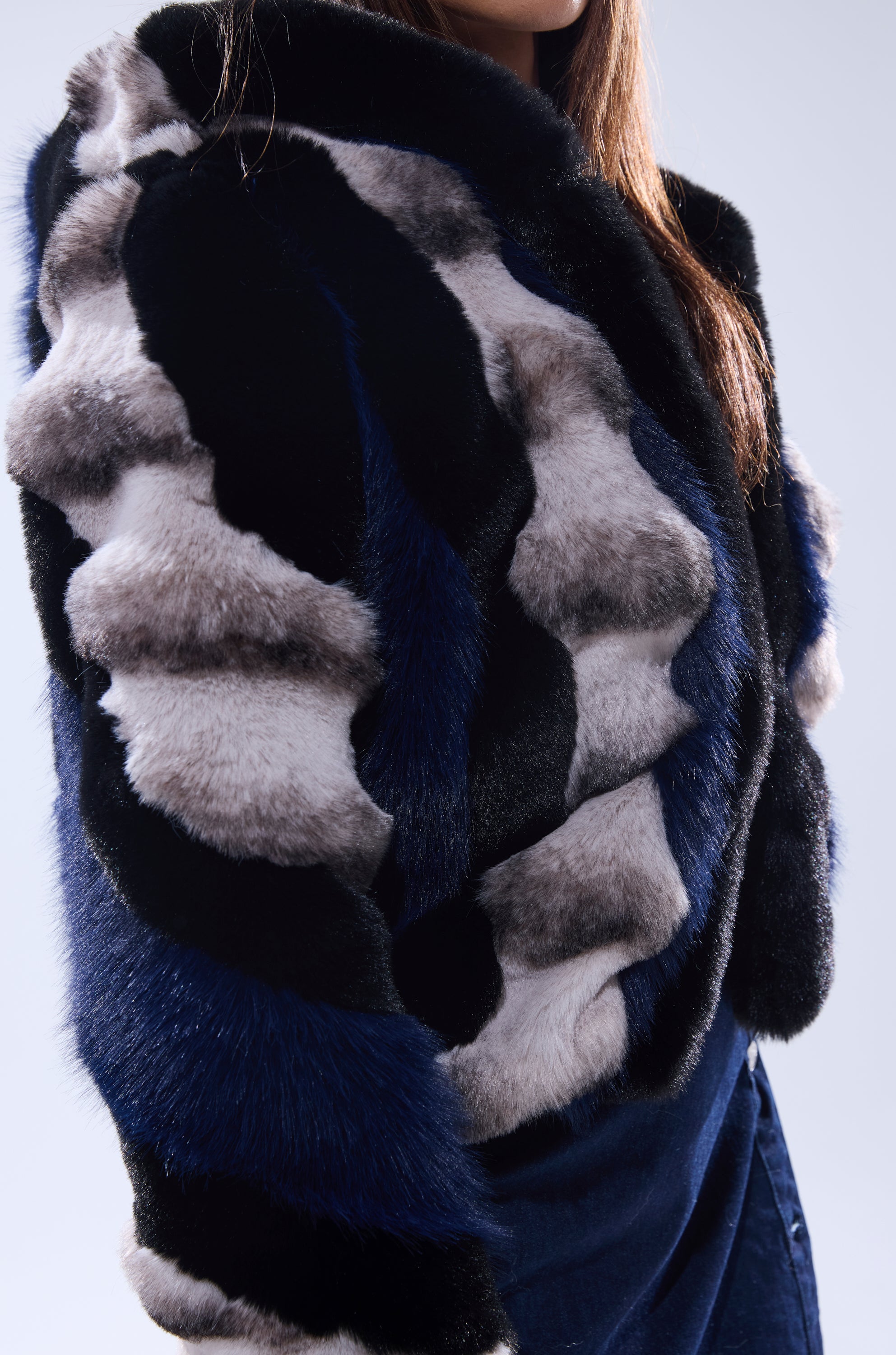A person wears the SUPERNOVA PANELED FAUX FUR IN BLUE MULTI, a luxurious patterned faux fur coat in black, blue, and gray shades, with long brown hair partially visible, standing against a light background.