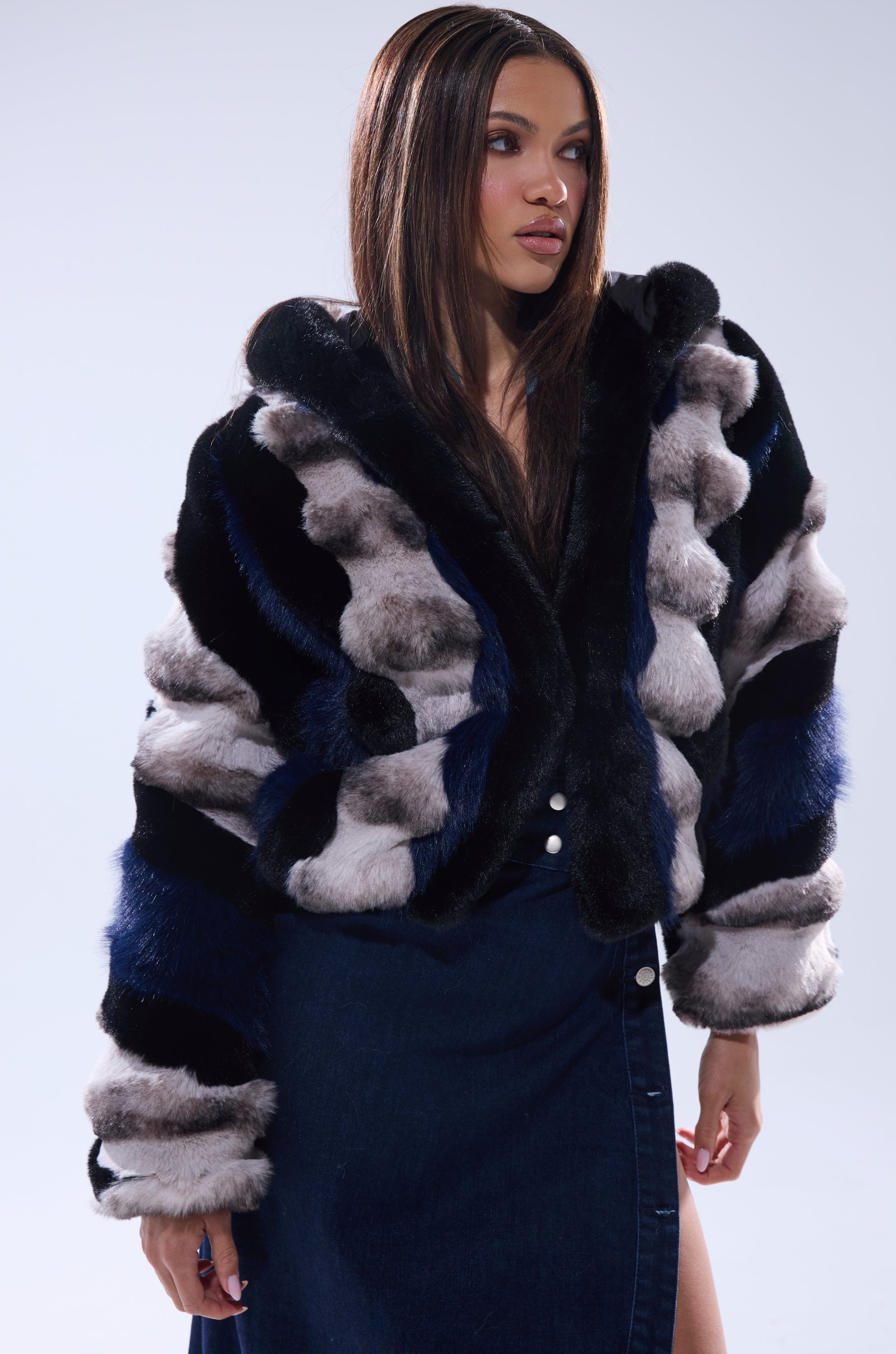 A woman models the SUPERNOVA PANELED FAUX FUR IN BLUE MULTI, paired with a dark blue front-slit skirt, standing against a light background and gazing confidently to the side.