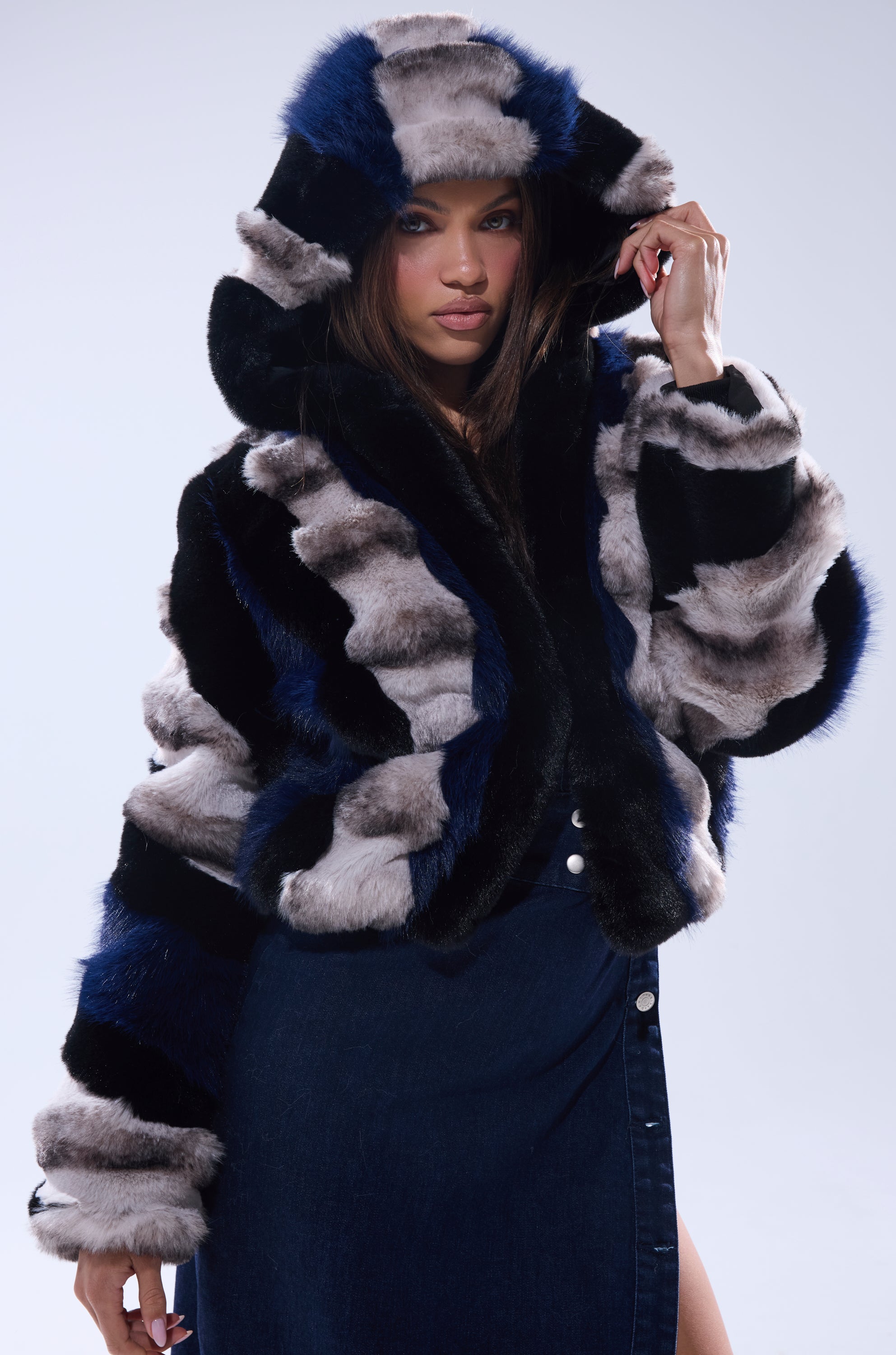 A woman wears the SUPERNOVA PANELED FAUX FUR IN BLUE MULTI jacket, holding the hood, paired with a dark blue denim skirt that has a front slit.