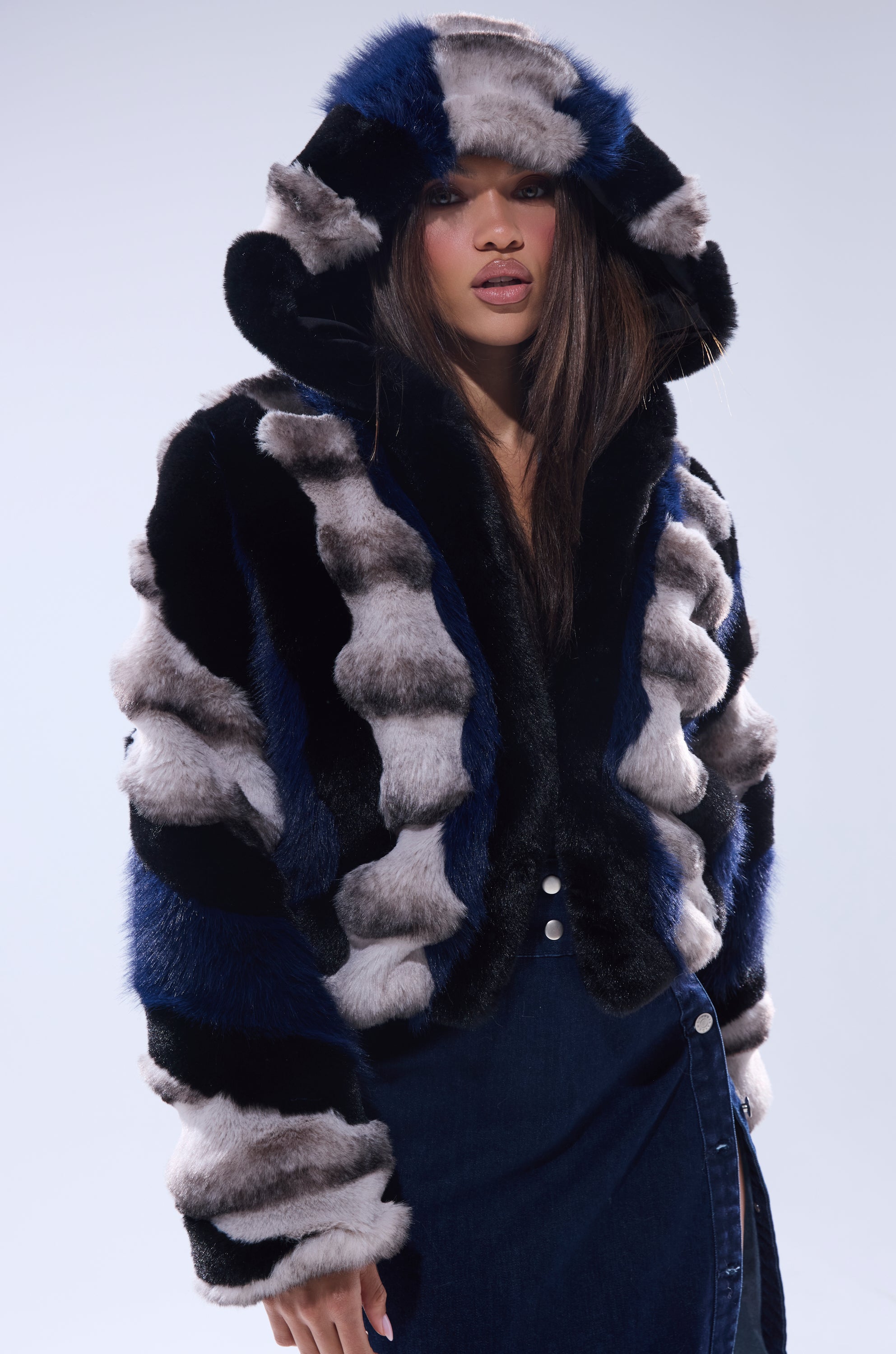 A woman in the SUPERNOVA PANELED FAUX FUR IN BLUE MULTI jacket, featuring bold black, blue, and gray stripes, stands against a plain light background. She has long straight hair and wears a dark denim buttoned skirt.