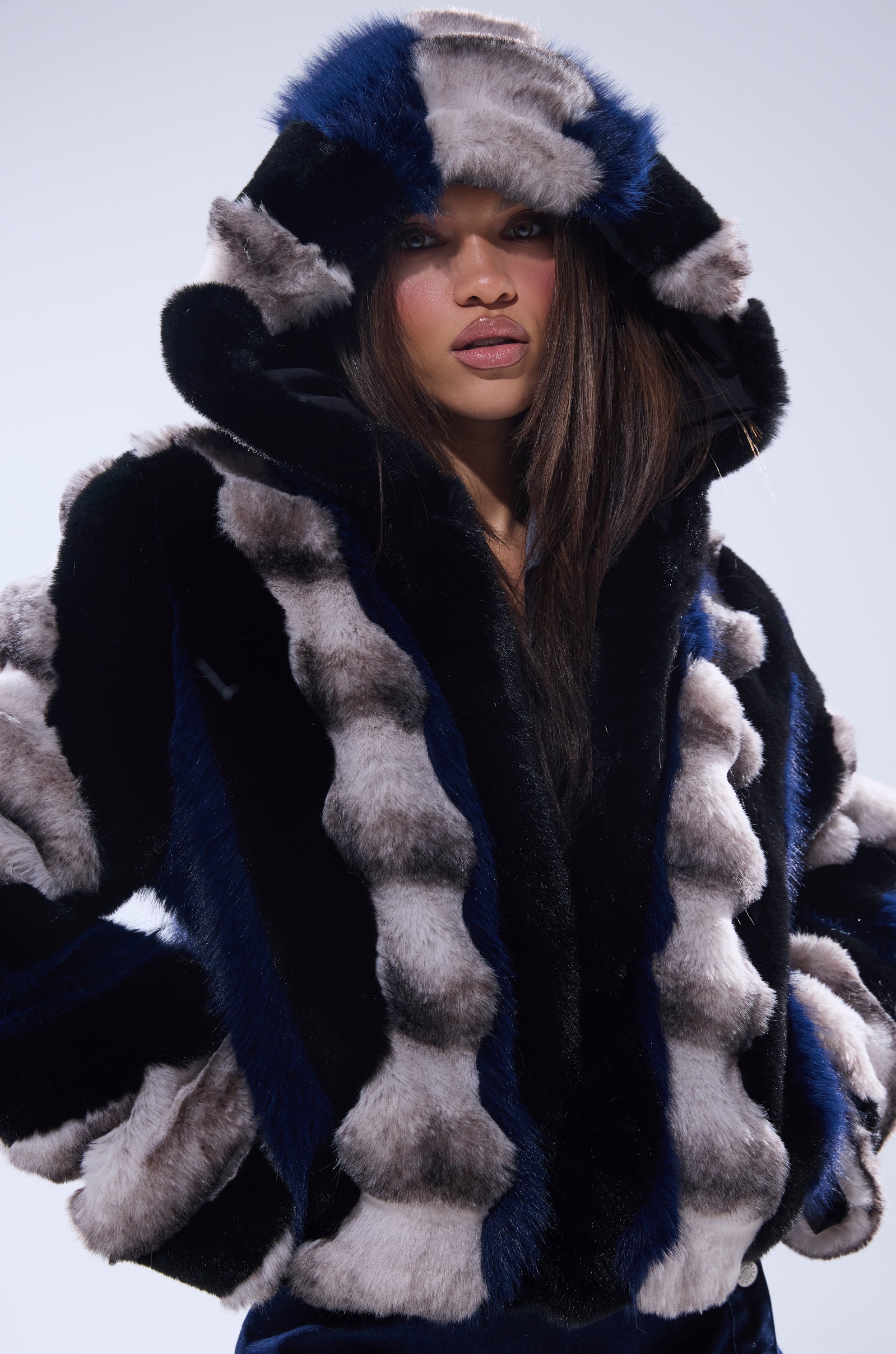 A woman wears the SUPERNOVA PANELED FAUX FUR IN BLUE MULTI, a hooded coat featuring bold black, gray, and deep blue stripes, as she stands confidently against a light background.