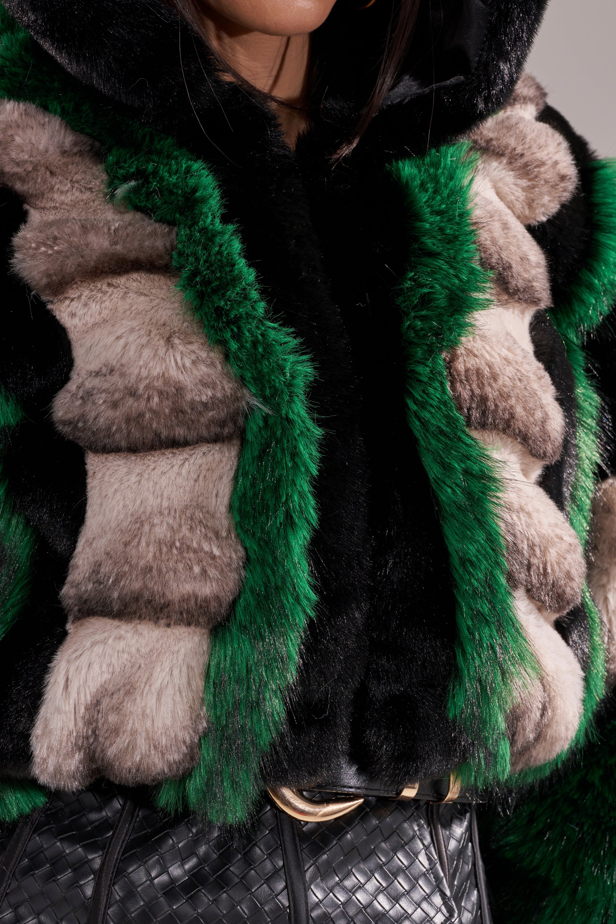 A person wears the SUPERNOVA Paneled Faux Fur in Green, featuring emerald green, black, and beige stripes with a black woven leather belt; only their lower face and upper body are visible.