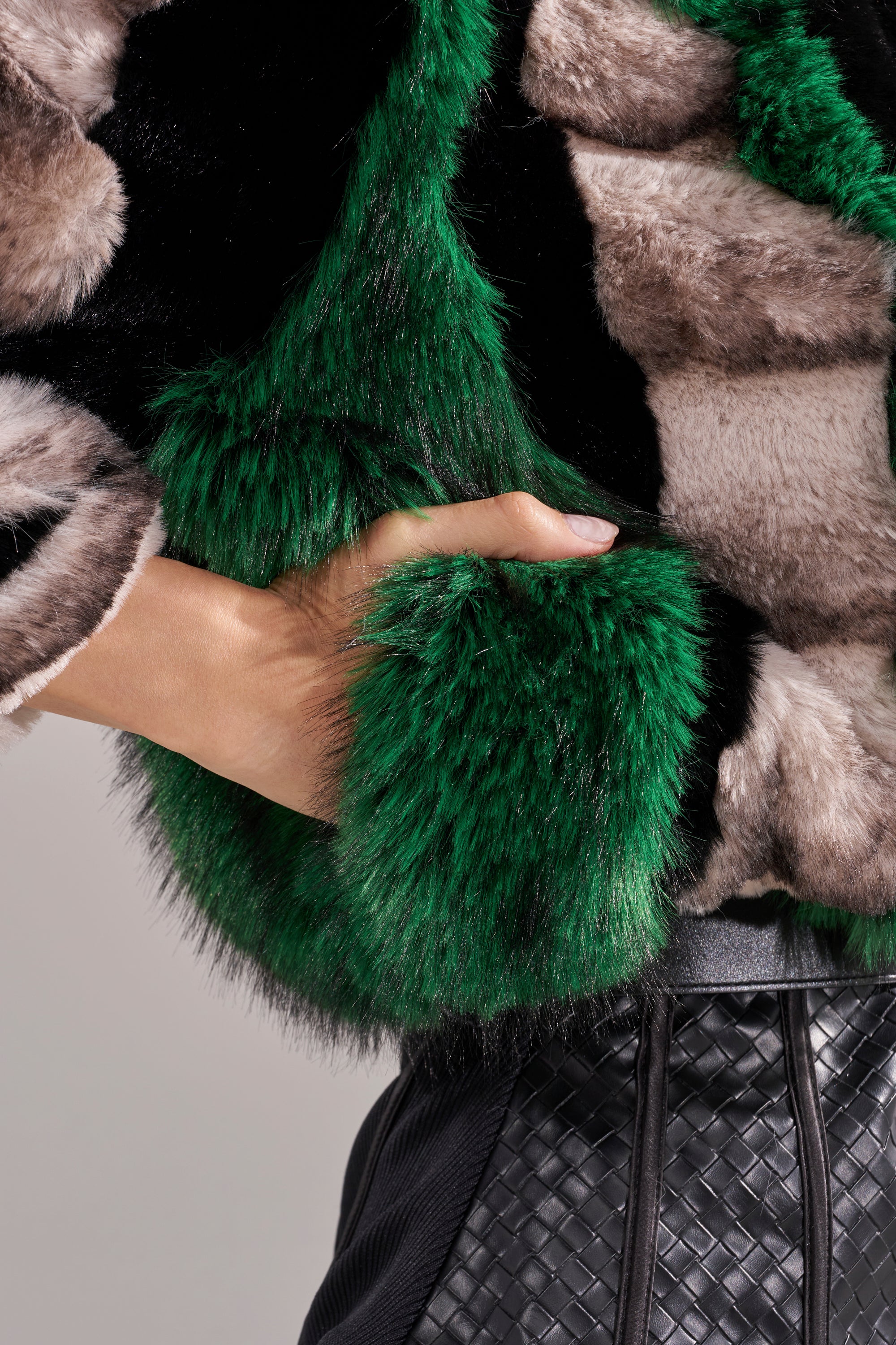 A close-up of a hand tucked into the pocket of the SUPERNOVA PANELED FAUX FUR IN GREEN coat, styled with black woven-texture pants.