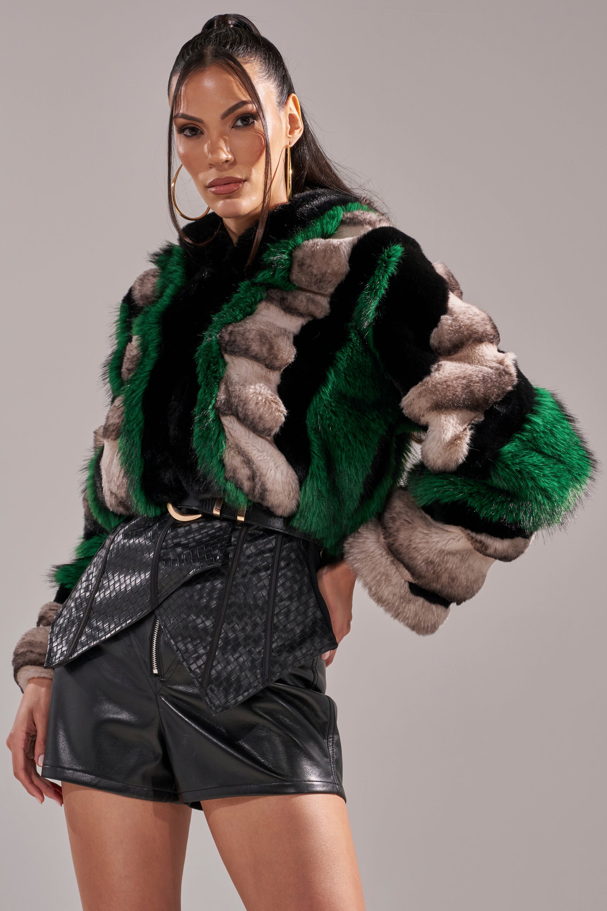 A woman poses confidently in the SUPERNOVA PANELED FAUX FUR IN GREEN, styled with high-waisted black leather shorts and hoop earrings, set against a plain gray background.