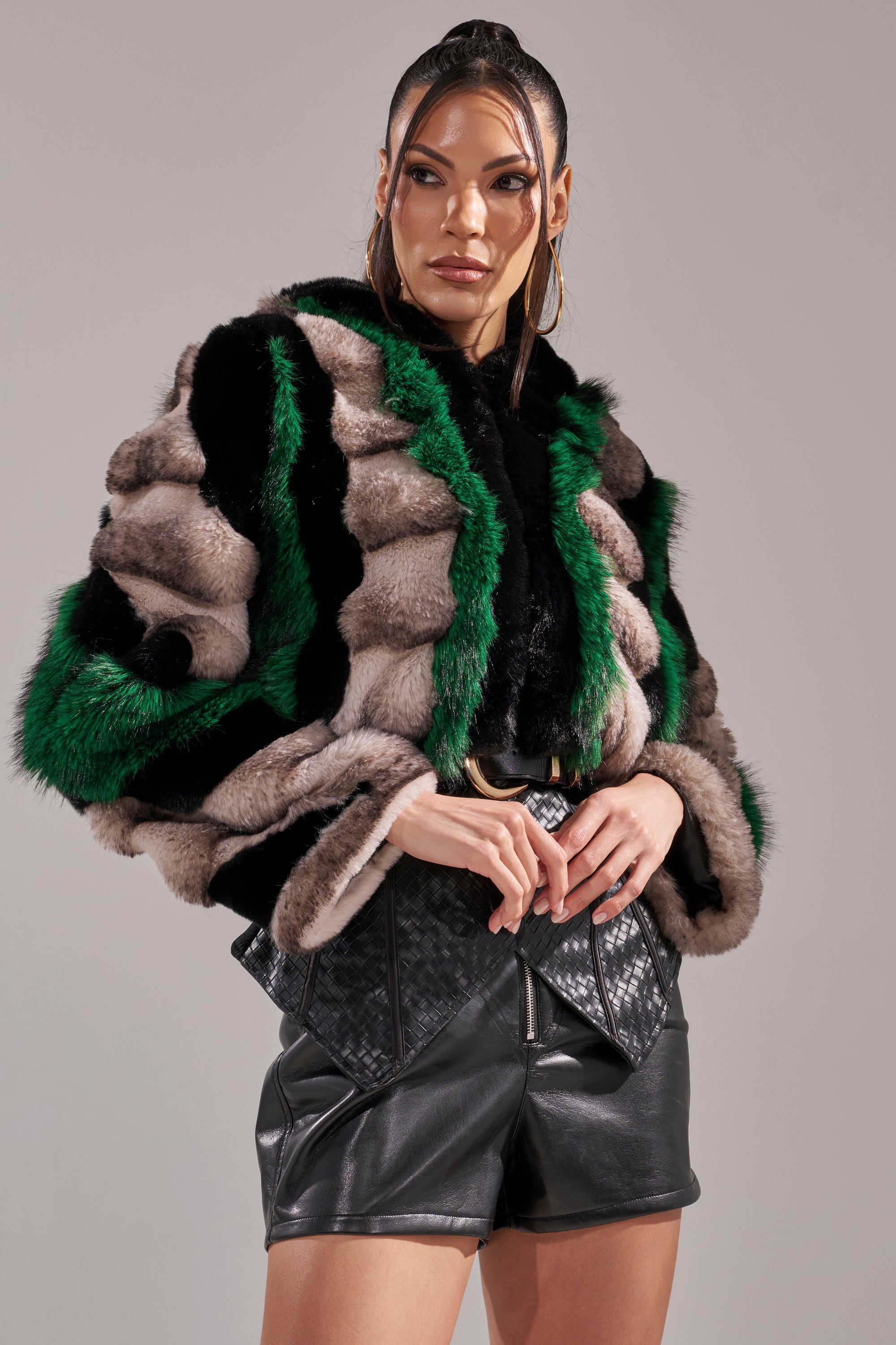 A woman with sleek, long hair confidently poses in the SUPERNOVA PANELED FAUX FUR IN GREEN, featuring bold green, black, and gray stripes, styled over high-waisted black leather shorts against a plain background.