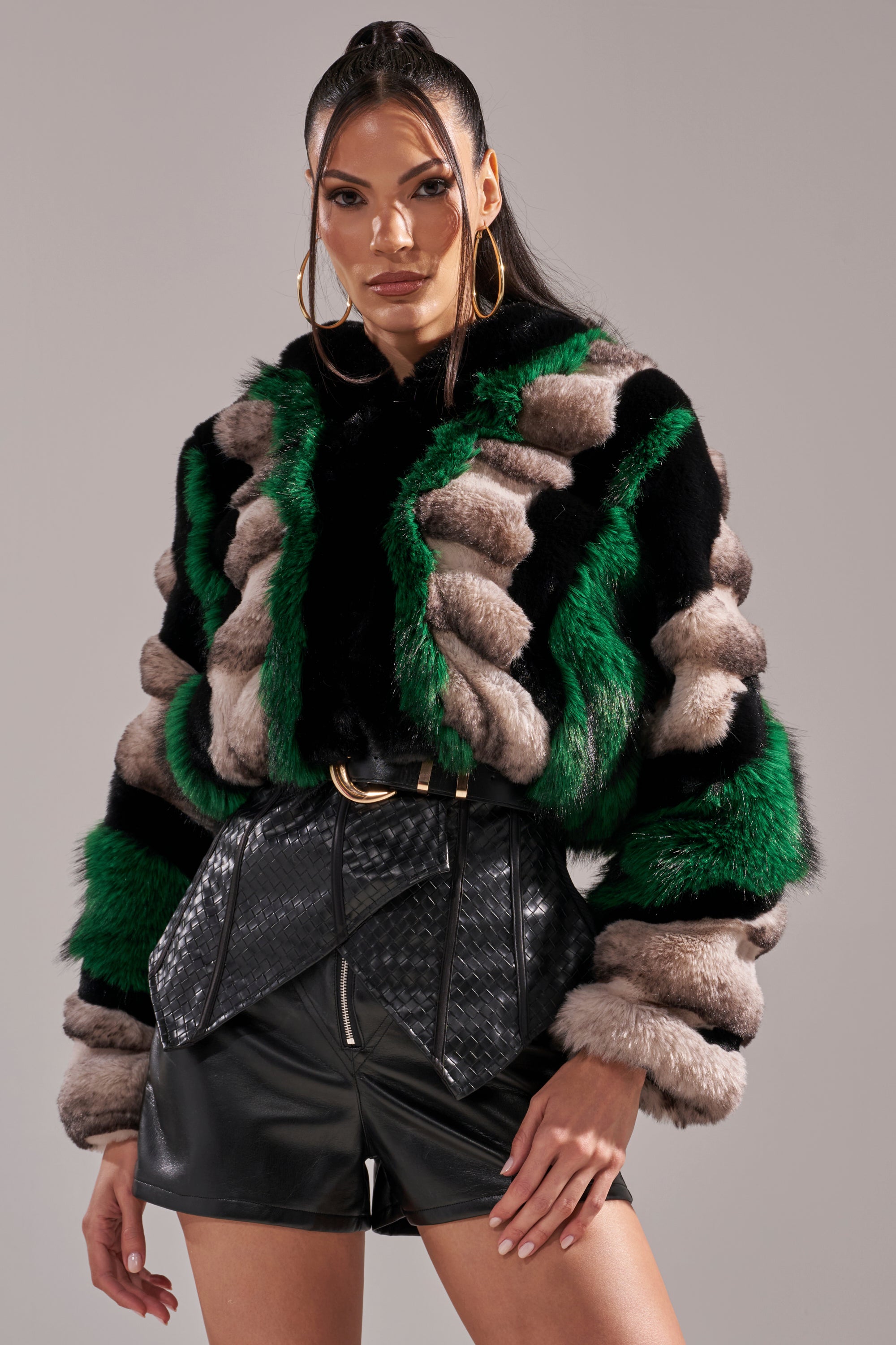 A woman stands confidently in the SUPERNOVA PANELED FAUX FUR IN GREEN, featuring black and beige stripes, styled with high-waisted black belted shorts, large gold hoop earrings, and a high ponytail.