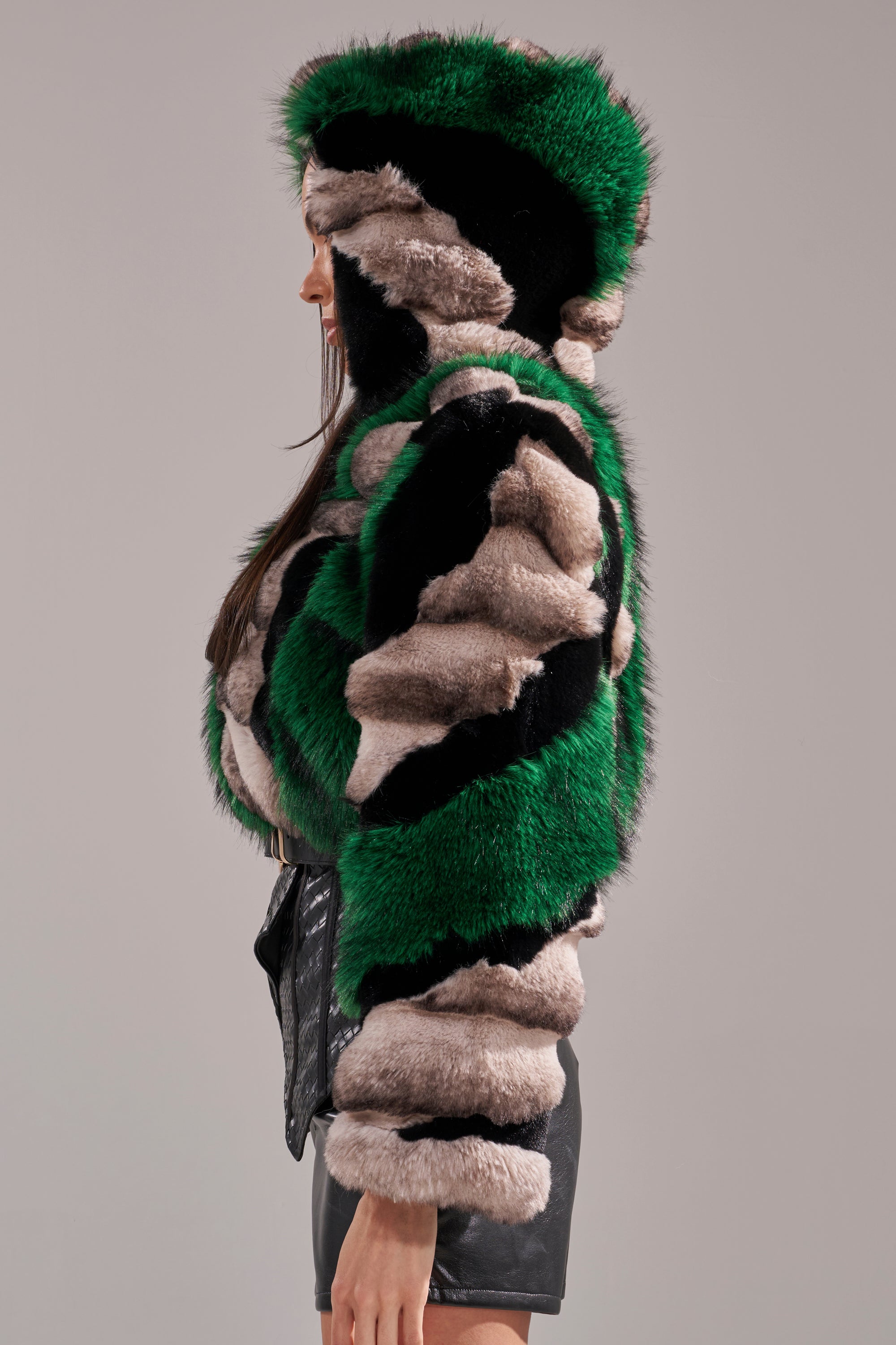 A person in profile wears the SUPERNOVA PANELED FAUX FUR IN GREEN, featuring green, black, and beige patches, paired with a black textured skirt against a plain gray background.