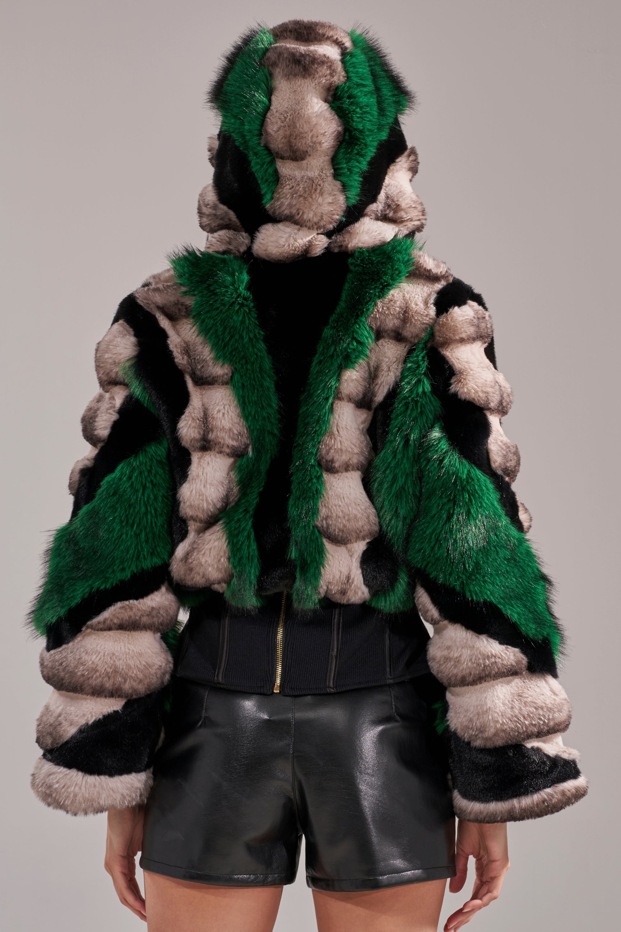 Someone is seen from the back against a plain background, wearing the SUPERNOVA PANELED FAUX FUR IN GREEN—a plush hooded coat with emerald green, black, and beige stripes—paired with shiny black leather shorts.