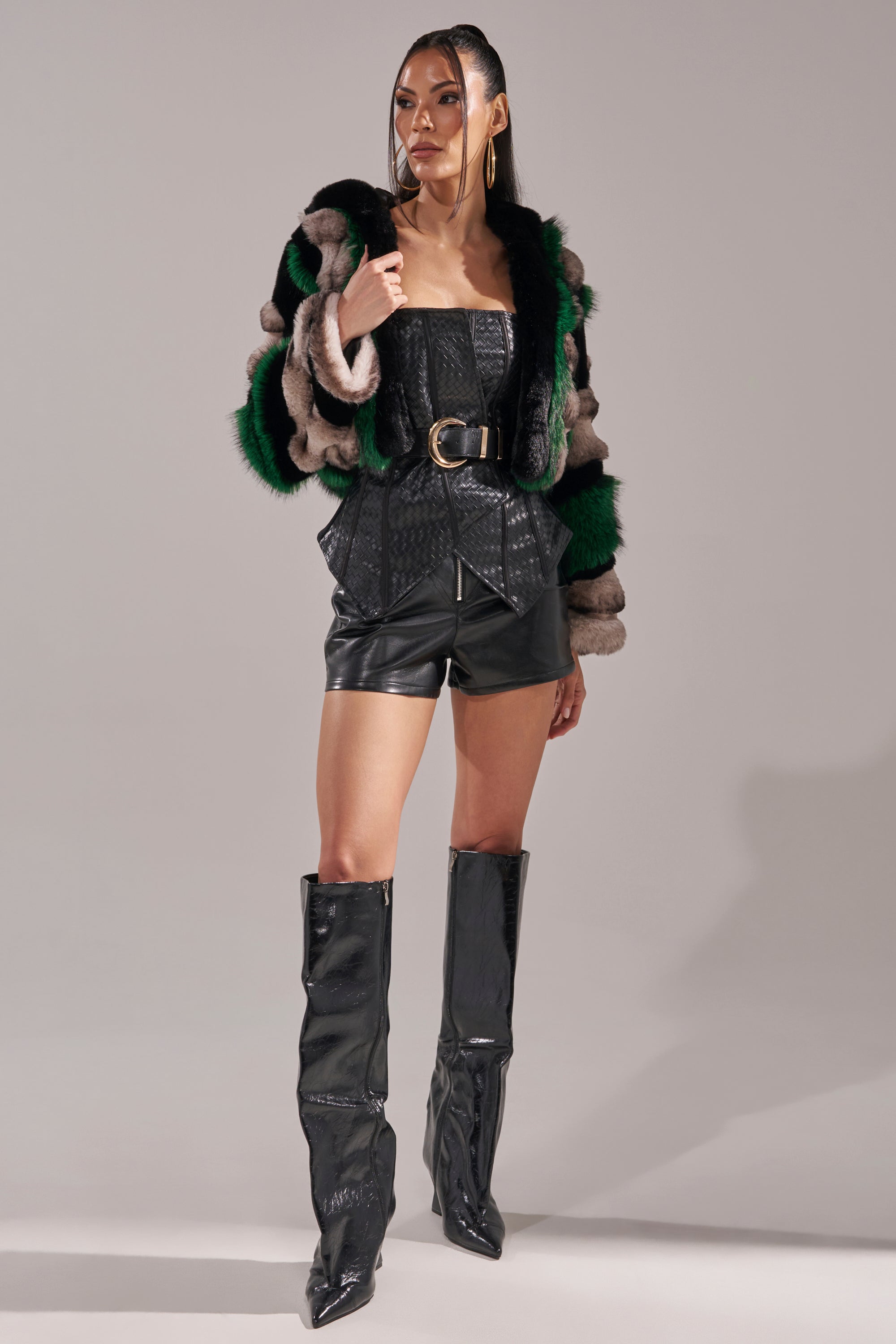 A woman models the SUPERNOVA PANELED FAUX FUR IN GREEN coat with brown and black accents, styled over a black faux leather belted top, matching shorts, and knee-high shiny black boots against a plain gray backdrop.