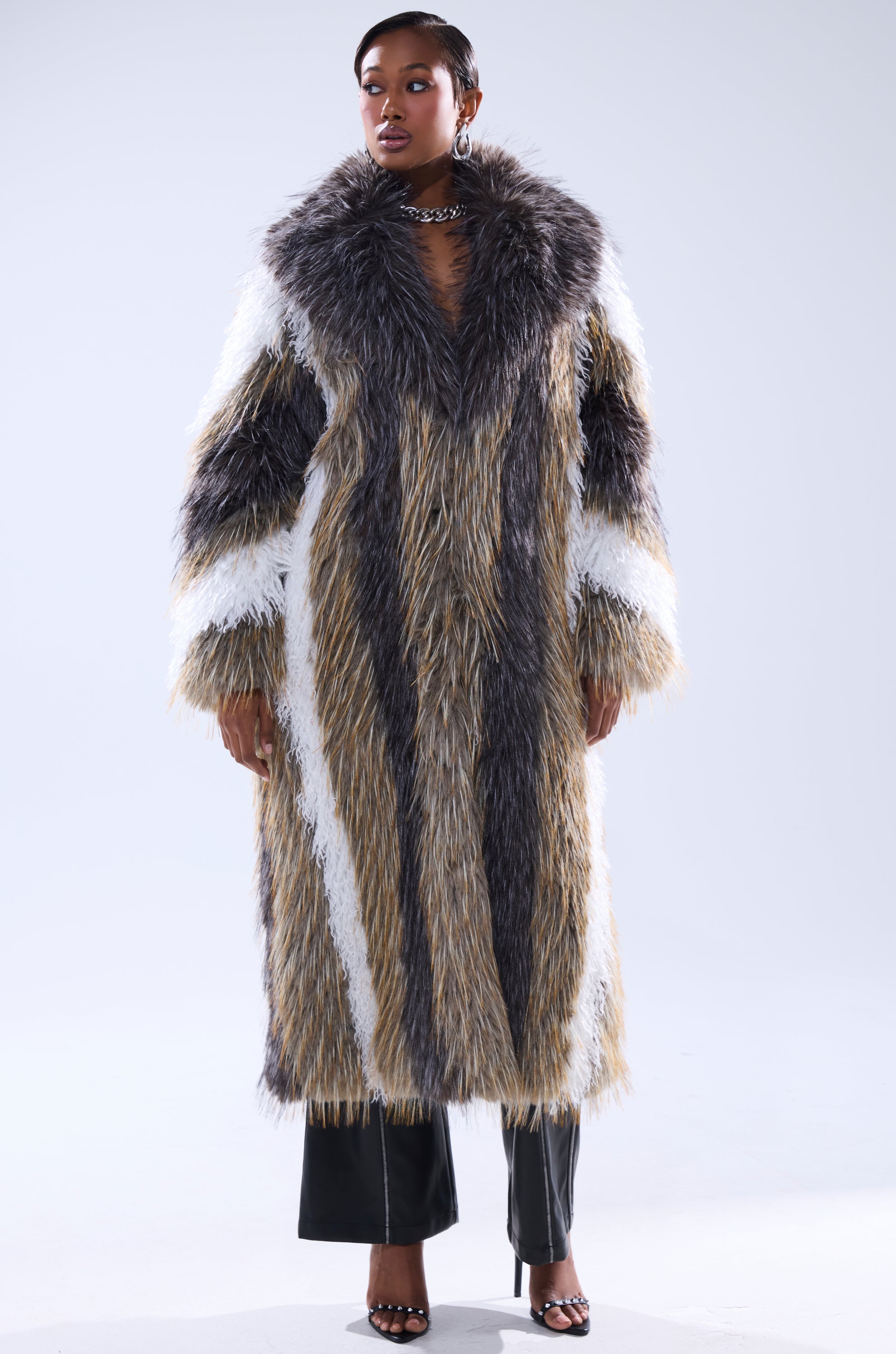 A woman faces forward against a plain light background, wearing the POISON IVY FAUX FUR COAT—an oversized, long coat with gray, white, and beige stripes—paired with black pants and open-toe heels.