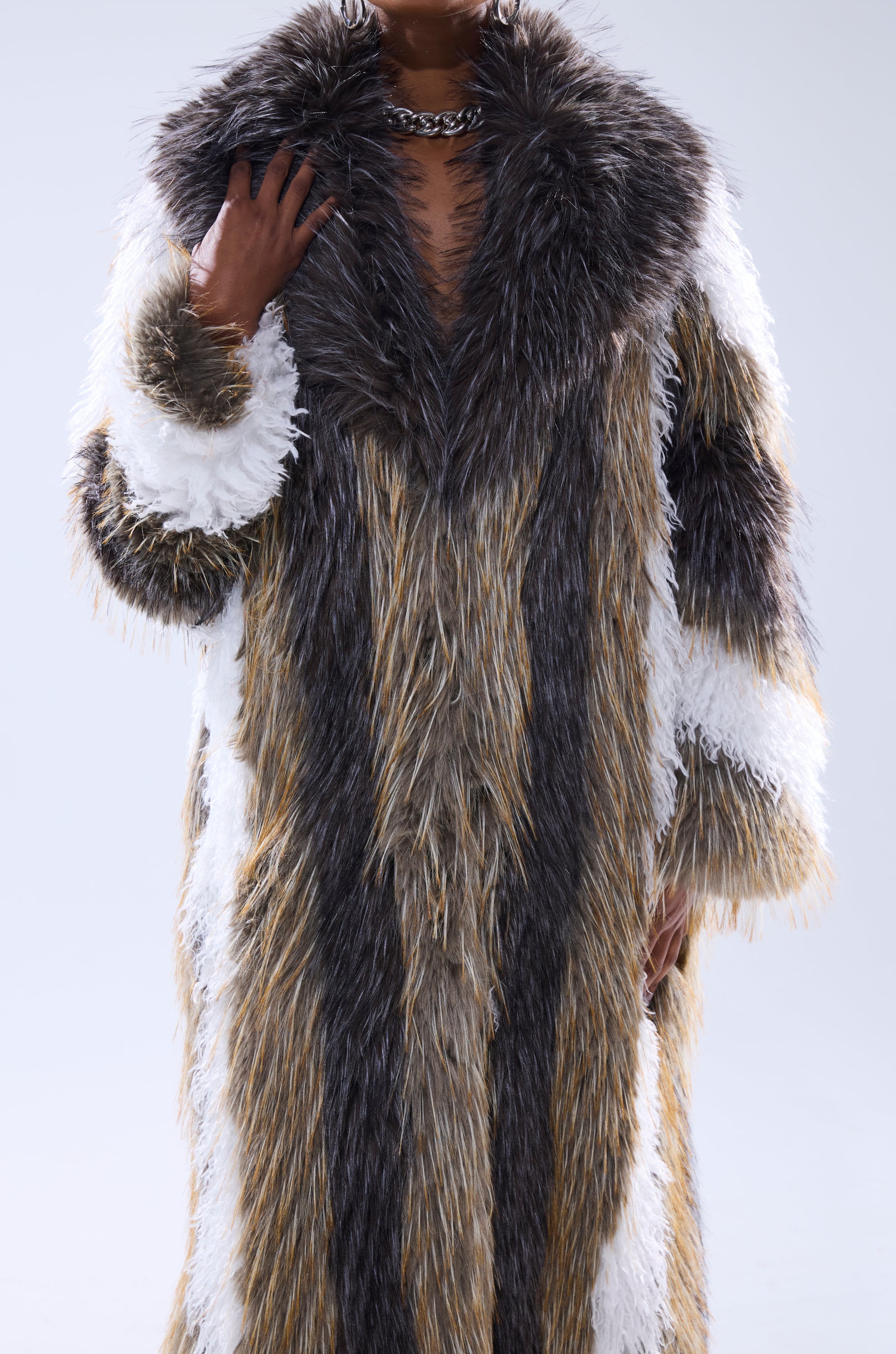 A person wears the POISON IVY FAUX FUR COAT, featuring long, shaggy faux fur in brown, gray, white, and black stripes, paired with a chunky silver chain necklace. The image is cropped at the neck and knees.