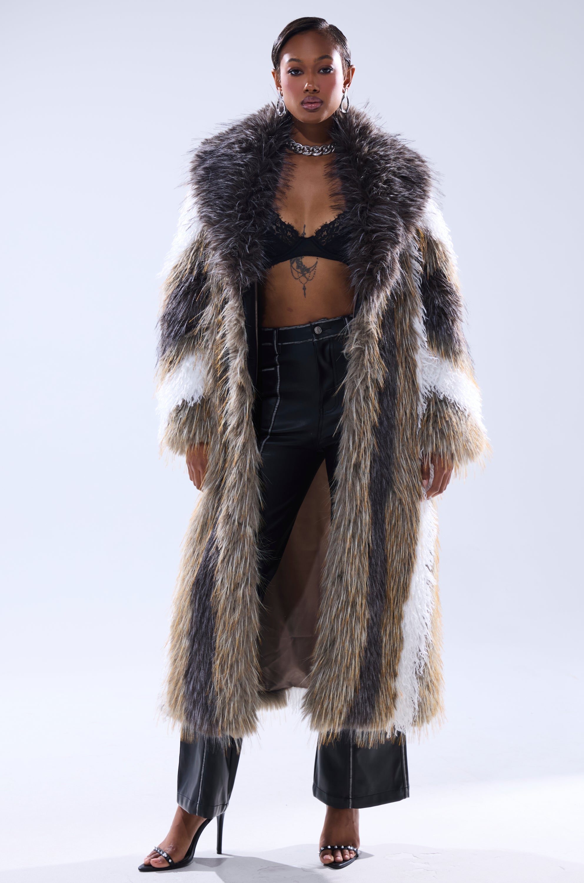 A woman stands confidently in the POISON IVY FAUX FUR COAT, its brown, white, and black stripes paired with a black lace bra, leather pants, open-toe heels, hoop earrings, and a chunky necklace.