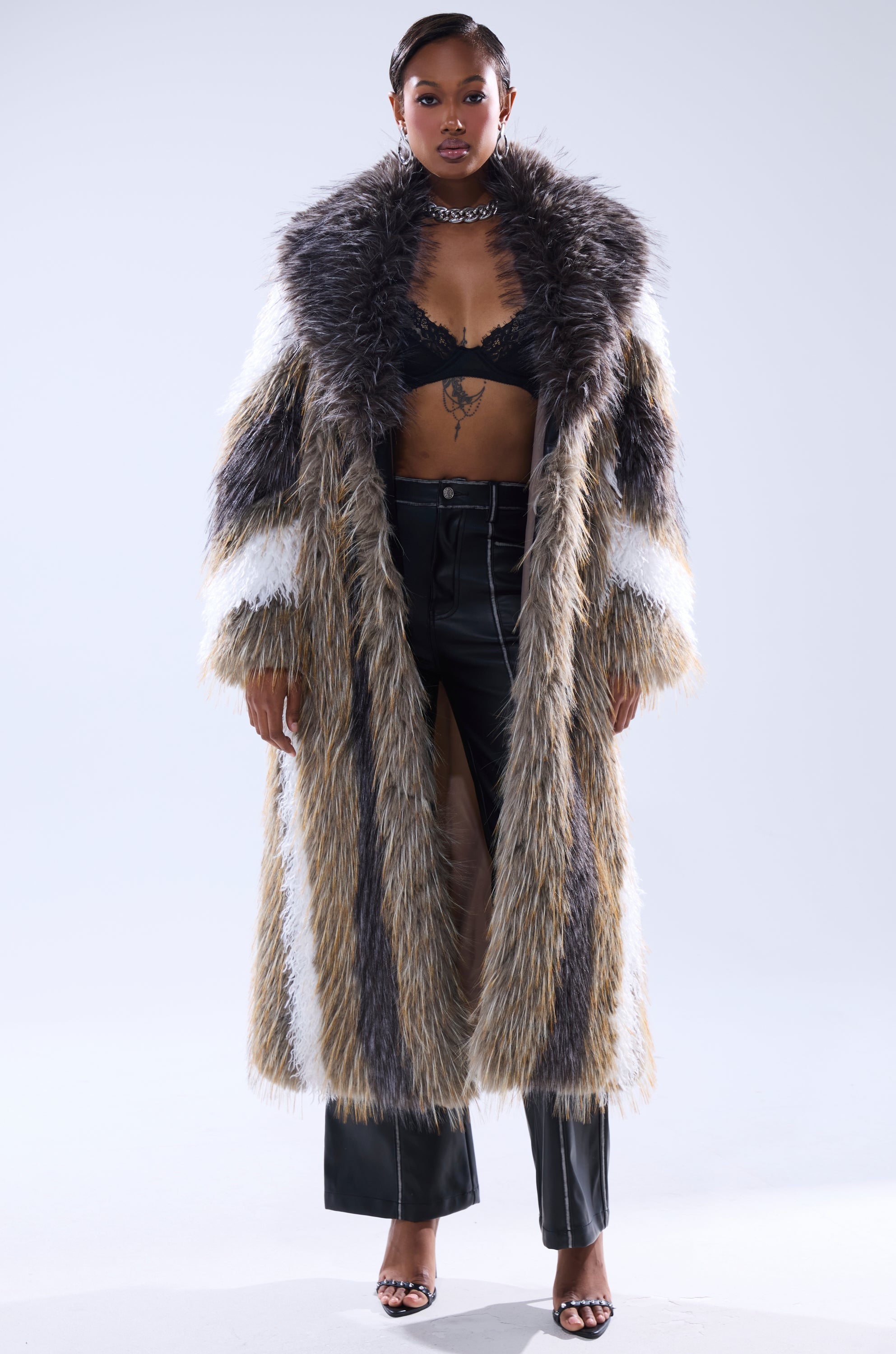 A woman stands confidently in the POISON IVY FAUX FUR COAT, layered over a black lace bralette, black leather pants, open-toe heels, and a chunky chain necklace against a plain background.